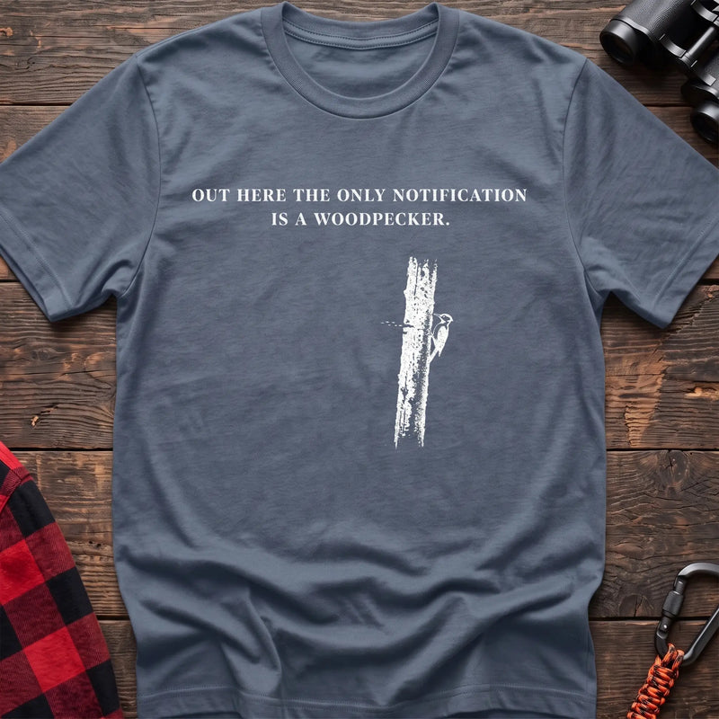 Stone blue cotton t-shirt with a woodpecker graphic and Out Here the Only Notification Is a Woodpecker text on rustic wood