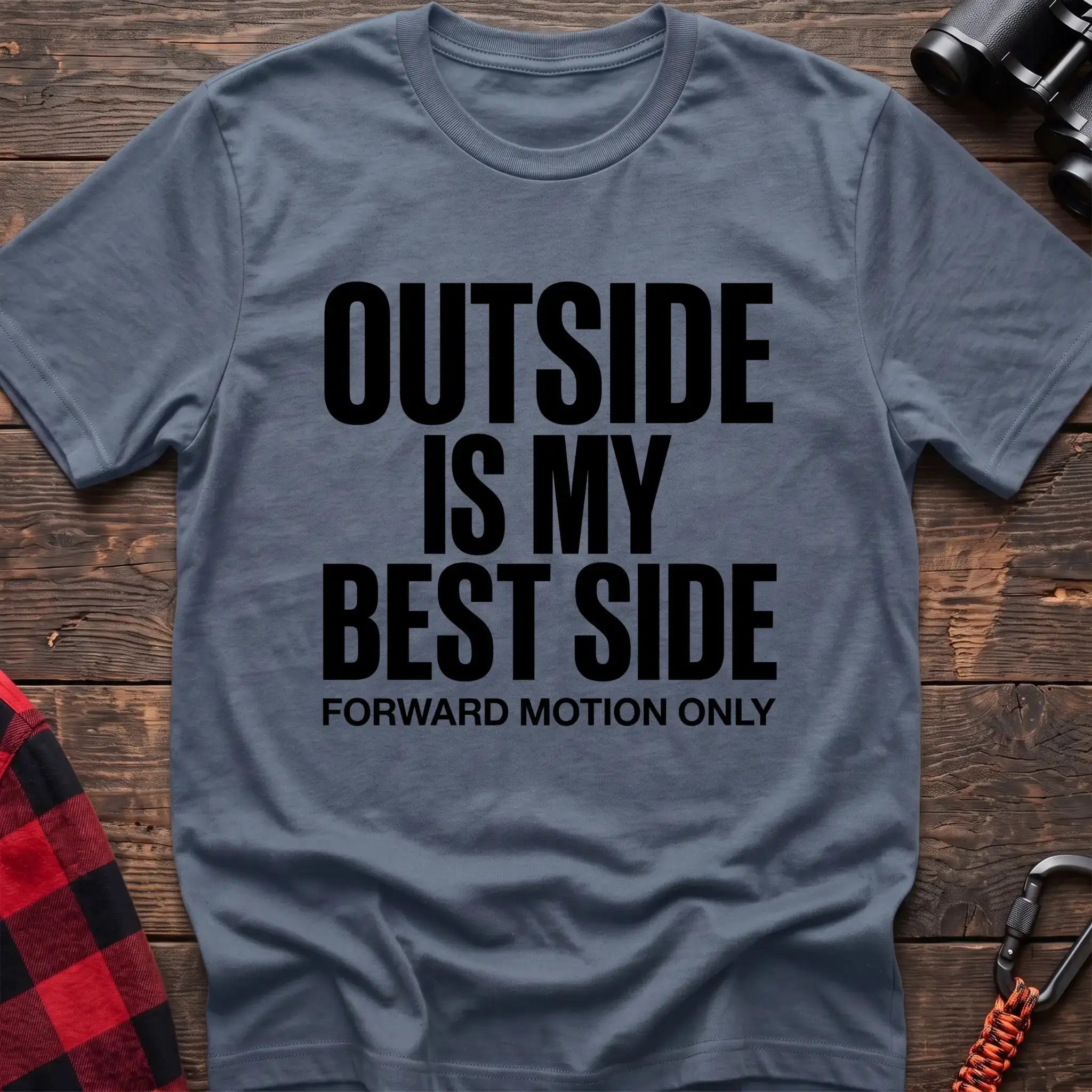Stone blue t-shirt with bold black text reading “OUTSIDE IS MY BEST SIDE” and “FORWARD MOTION ONLY” below it, laid flat on a wooden surface.