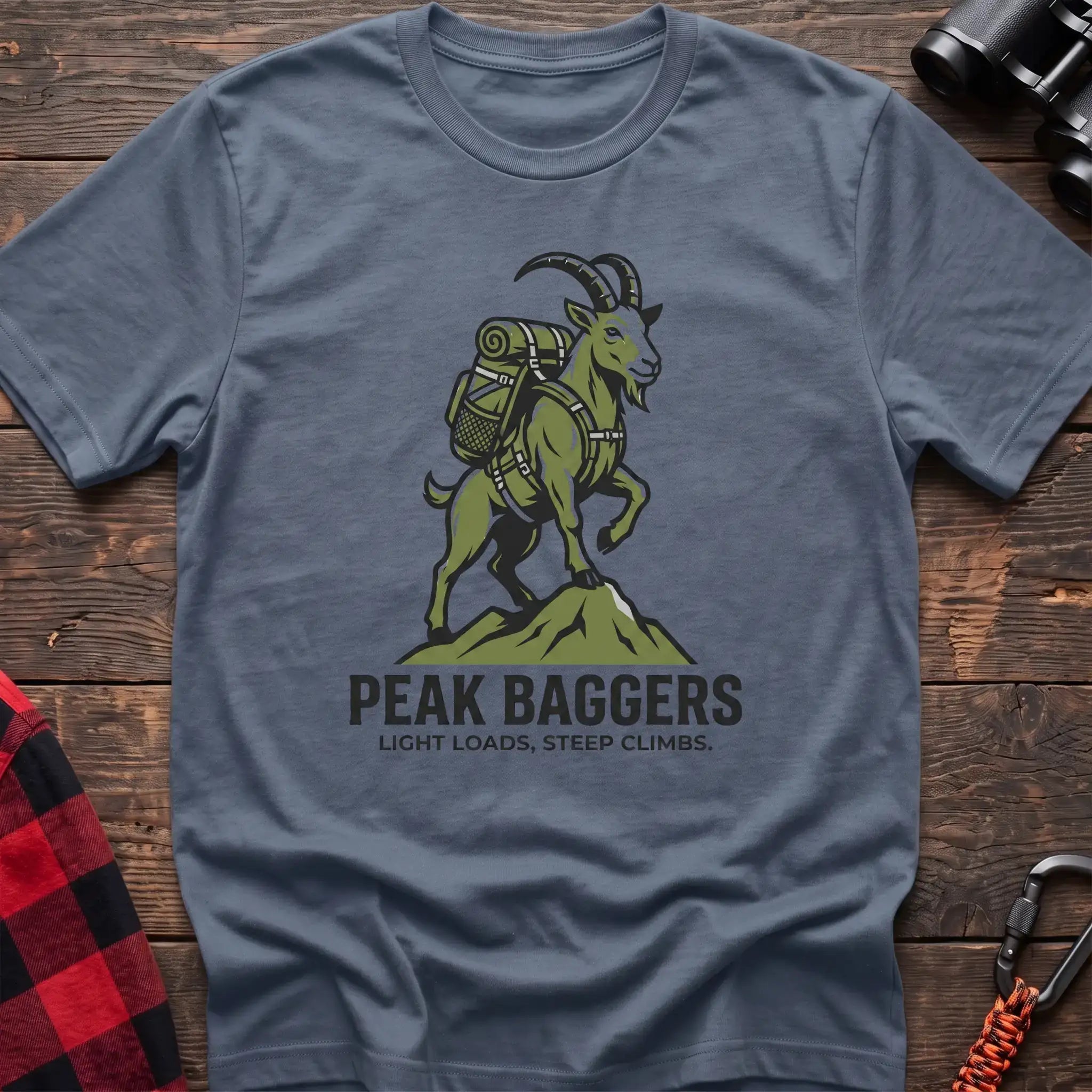 Stone blue t-shirt with an illustration of a mountain goat wearing a backpack standing on a peak, with the text “PEAK BAGGERS” and “LIGHT LOADS, STEEP CLIMBS,” laid flat on a wooden surface.