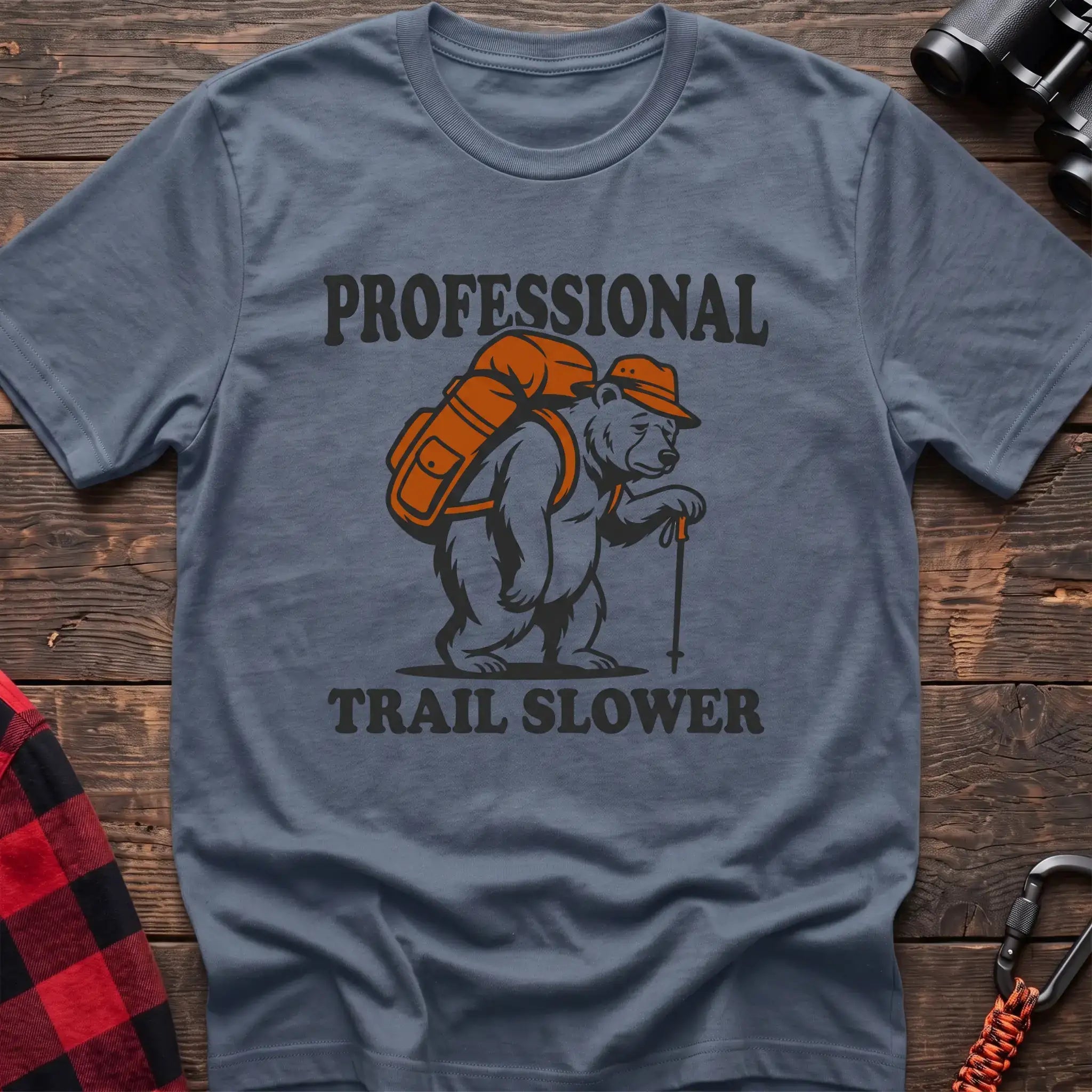 Stone blue t-shirt with a cartoon polar bear wearing a backpack and hat while holding a hiking stick, with the text “PROFESSIONAL TRAIL SLOWER,” laid flat on a wooden surface.
