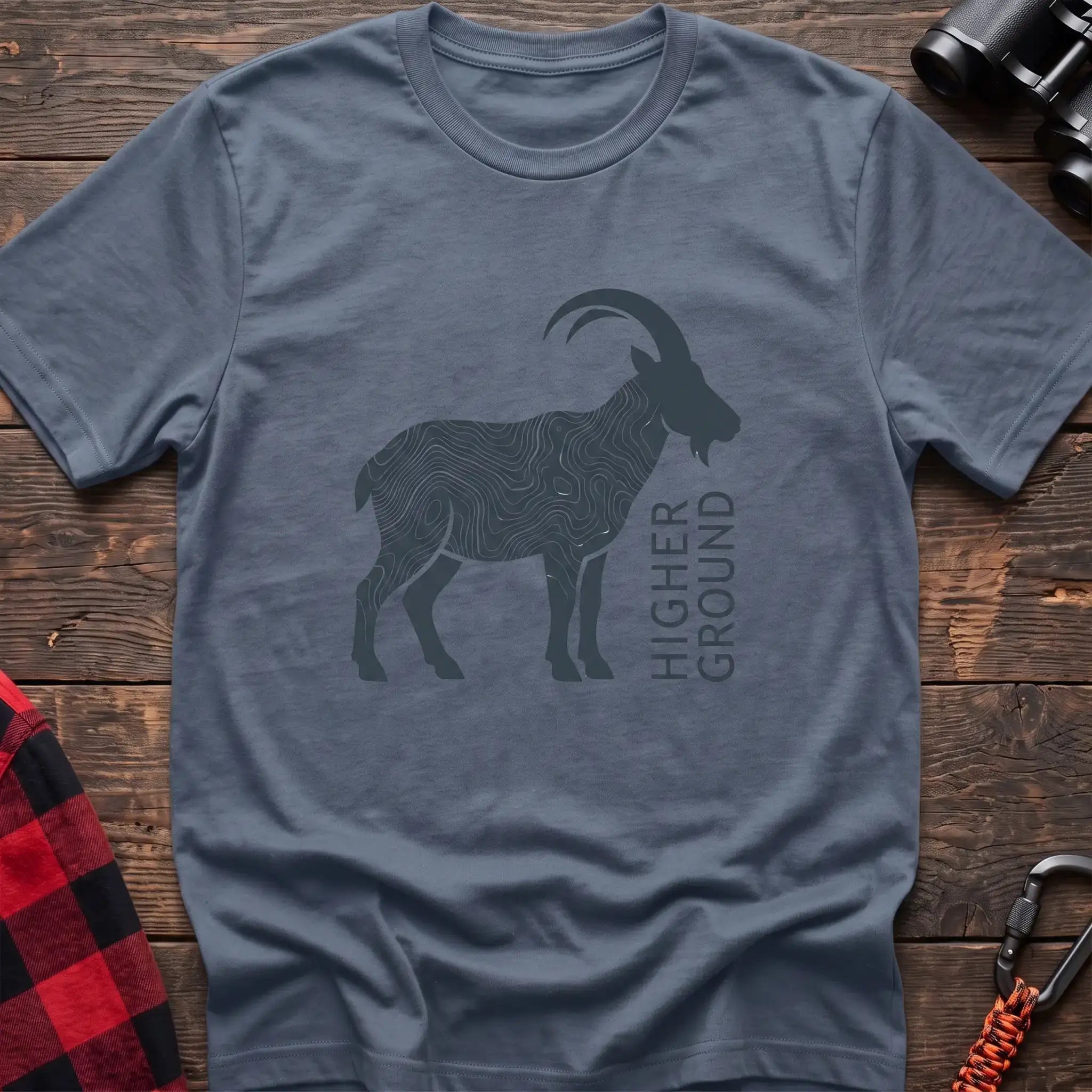 Stone blue t-shirt with a dark silhouette of a mountain goat filled with topographic contour lines, and the vertical text “HIGHER GROUND” printed beside it, laid flat on a wooden surface.