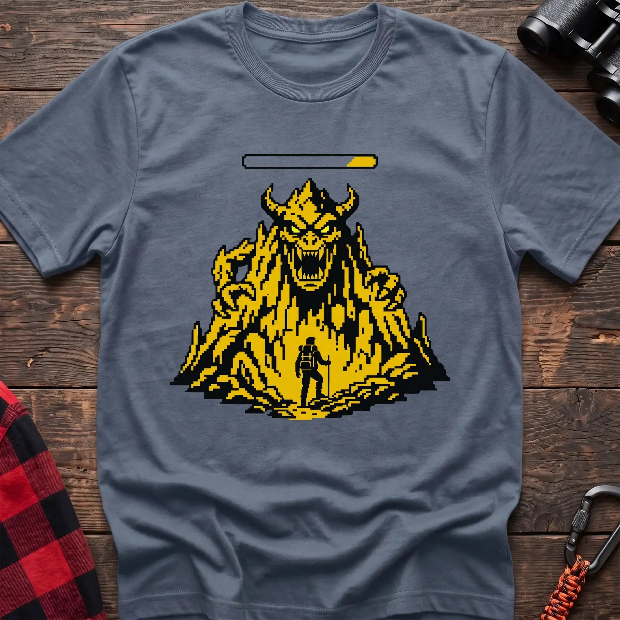 Stone blue t-shirt with a pixel-art illustration of a hiker facing a large horned mountain monster with a health bar above it, laid flat on a wooden surface.