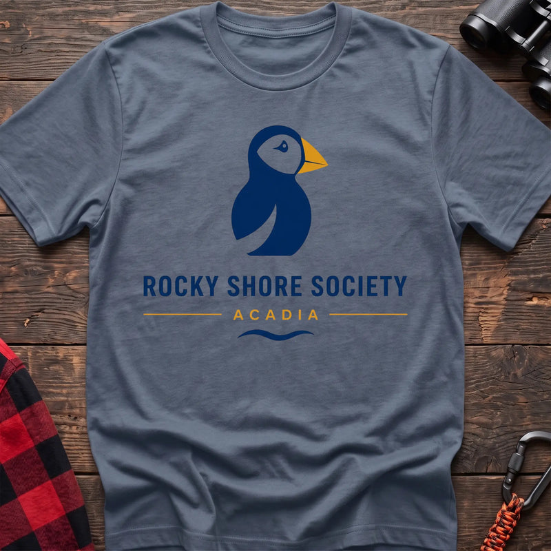 Stone blue cotton t-shirt with a navy puffin logo, yellow beak, and Rocky Shore Society Acadia text on a rustic wood background