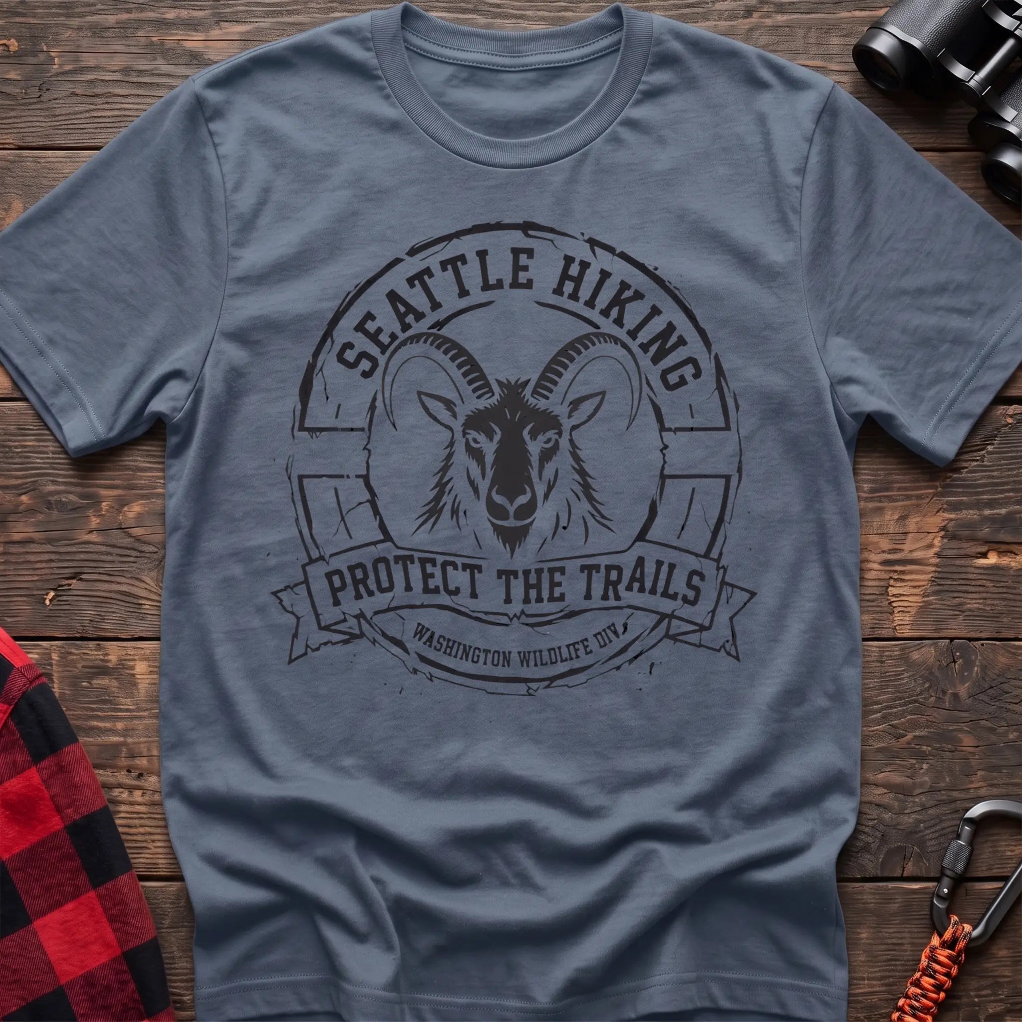 Stone blue cotton t-shirt with vintage Seattle Hiking graphic of a mountain goat in a circular badge, Protect the Trails Washington Wildlife Division design, flat lay on rustic wood with hiking gear