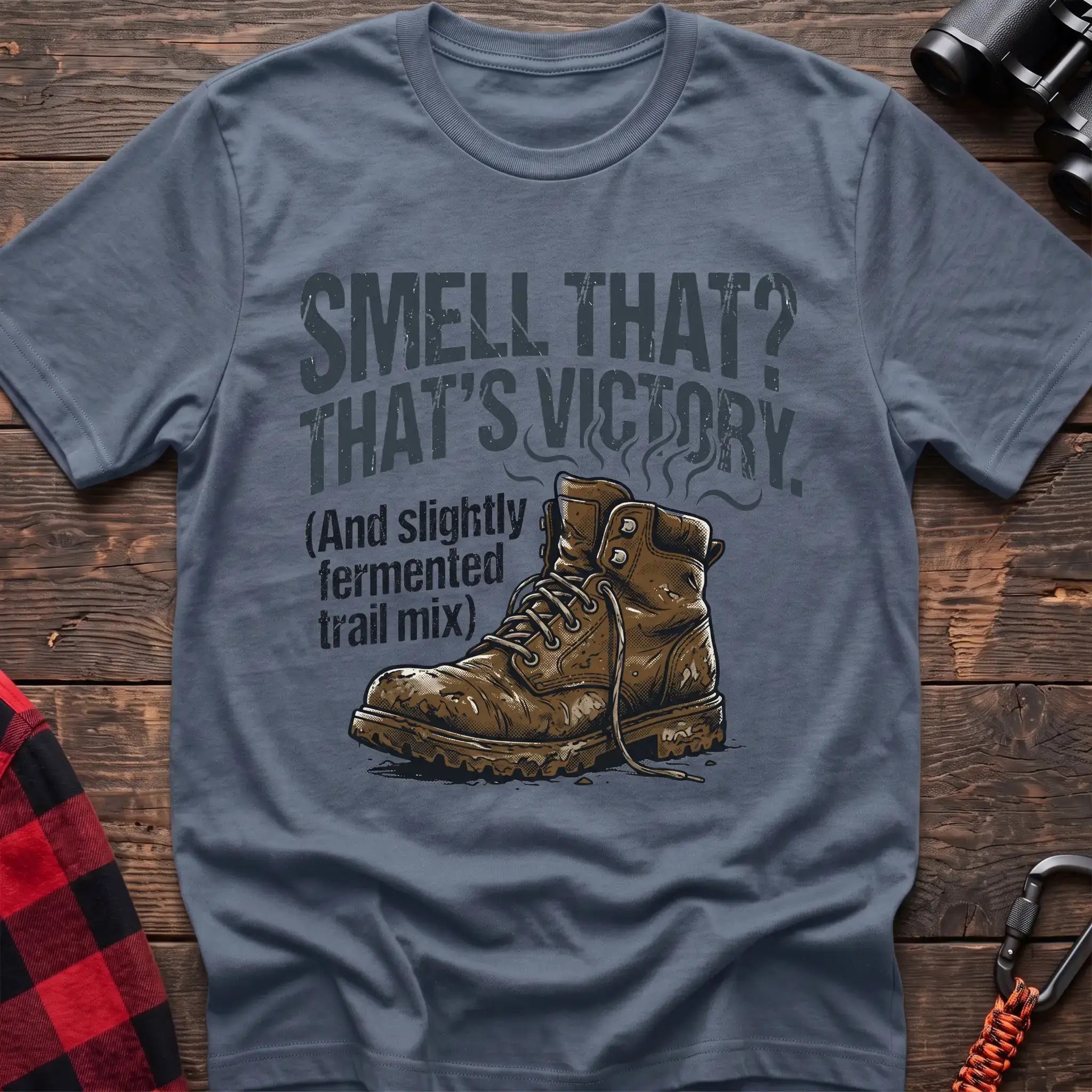 Stone blue t-shirt with an illustration of a dirty hiking boot with rising steam lines, and the text “SMELL THAT? THAT’S VICTORY. (And slightly fermented trail mix),” laid flat on a wooden surface