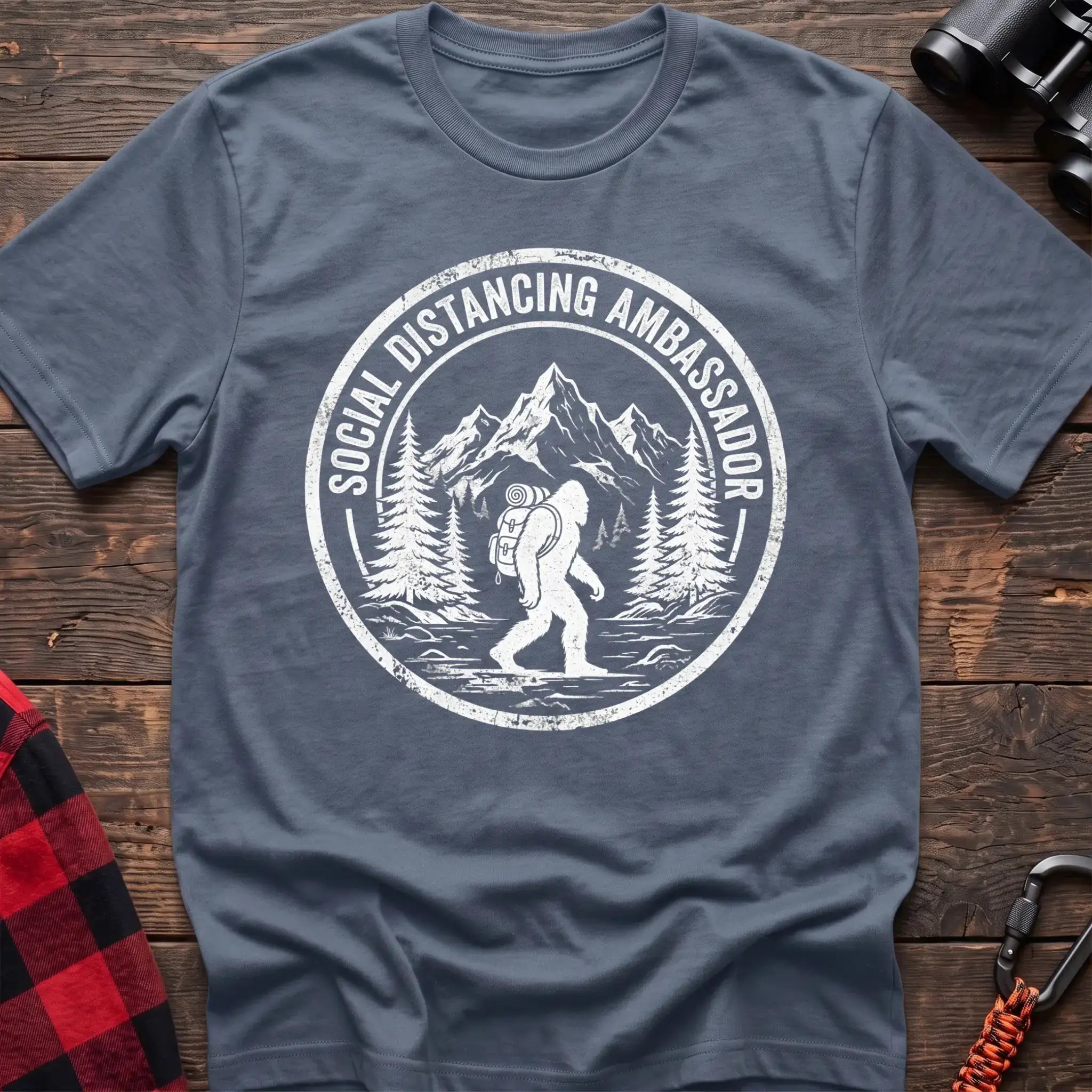 Stone blue t-shirt with white circular badge graphic of Bigfoot hiking with a backpack through mountains and the text Social Distancing Ambassador
