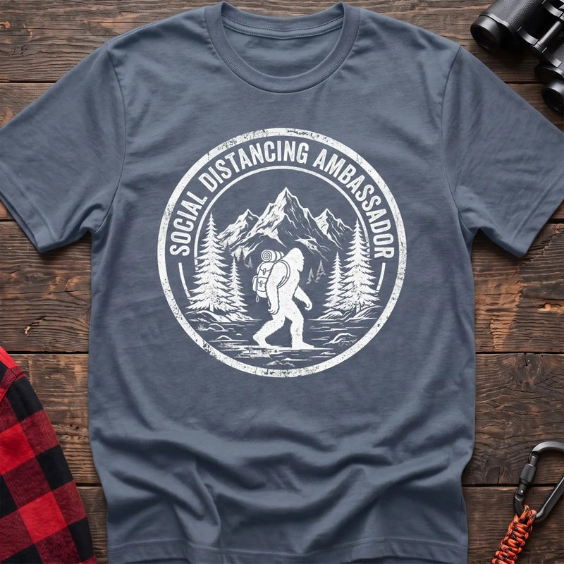 Stone blue t-shirt with white circular badge graphic of Bigfoot hiking with a backpack through mountains and the text Social Distancing Ambassador
