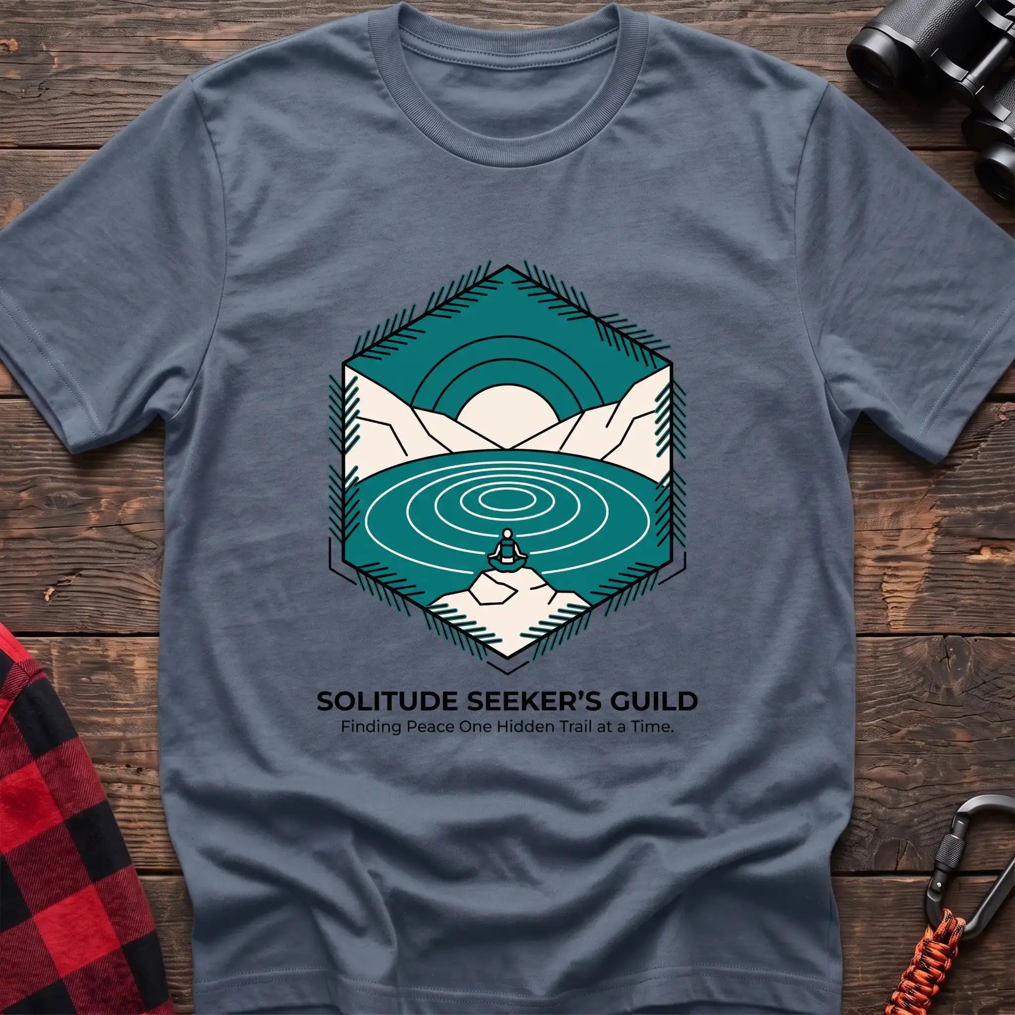 Stone blue t-shirt with a hexagonal illustration of a person meditating by a mountain lake, with the text “SOLITUDE SEEKER’S GUILD” and “Finding Peace One Hidden Trail at a Time,” laid flat on a wooden surface.