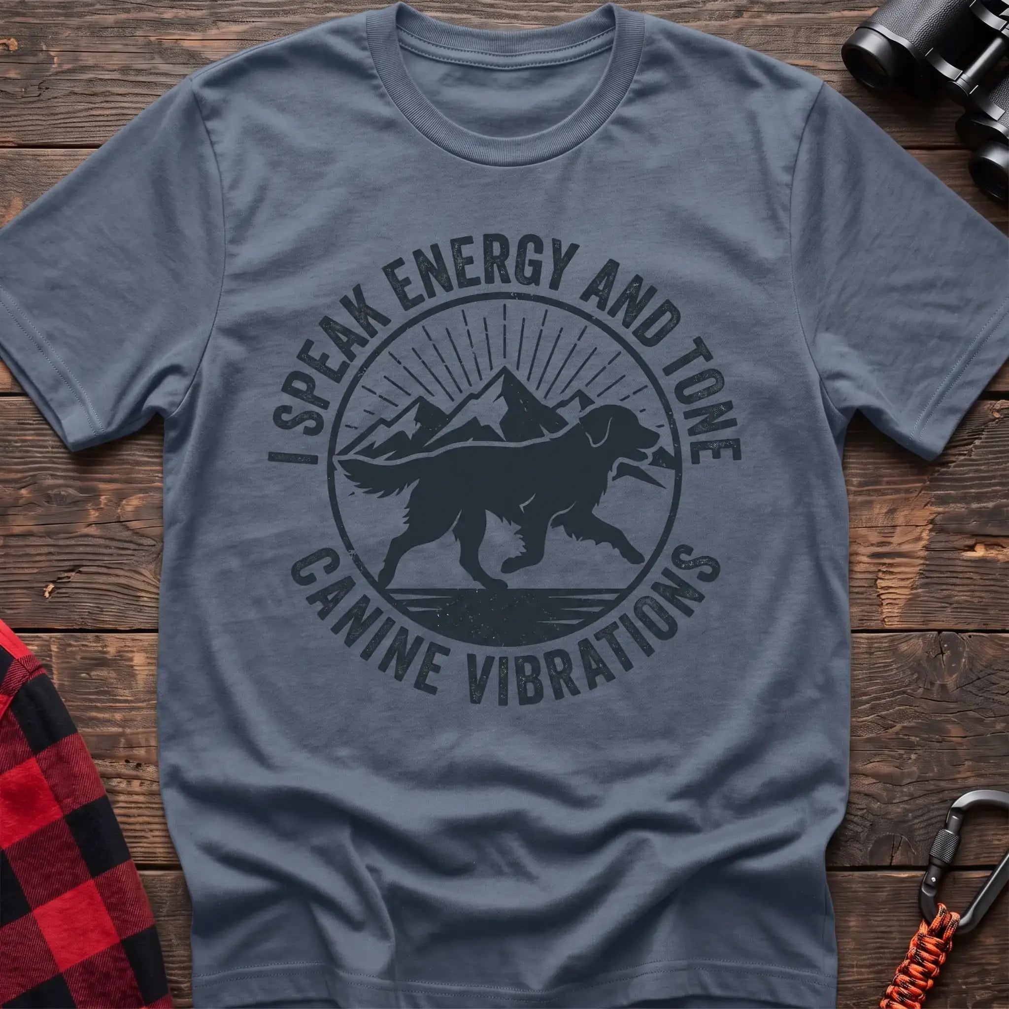 Stone blue t-shirt with a circular dark gray badge design featuring a running dog carrying a stick against mountain peaks and sun rays, with the text “I SPEAK ENERGY AND TONE” and “CANINE VIBRATIONS,” laid flat on a wooden surface.