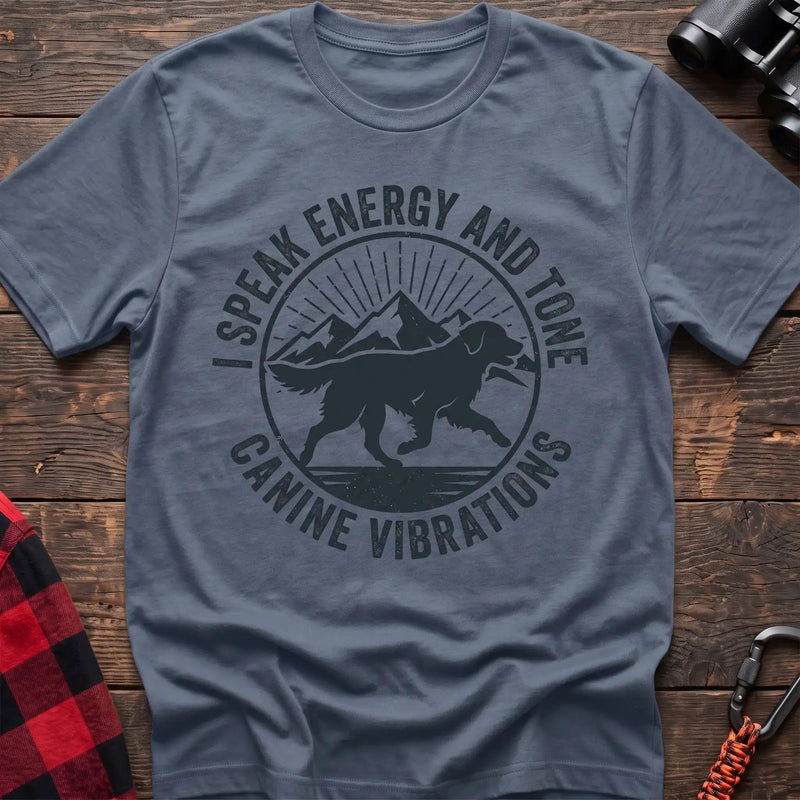 Stone blue t-shirt with a circular dark gray badge design featuring a running dog carrying a stick against mountain peaks and sun rays, with the text “I SPEAK ENERGY AND TONE” and “CANINE VIBRATIONS,” laid flat on a wooden surface.