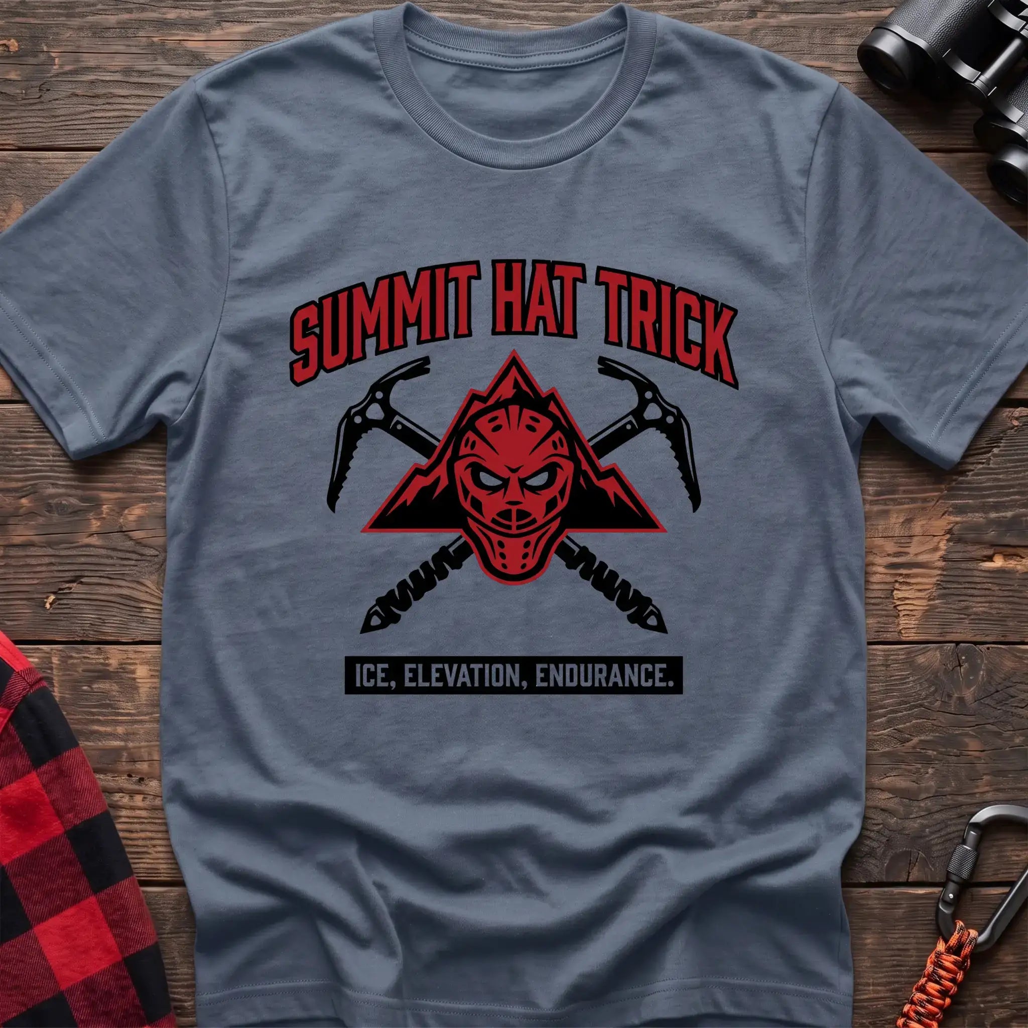 Stone blue t-shirt with a red mountain-themed goalie mask and crossed ice axes, with the text “SUMMIT HAT TRICK” and “ICE, ELEVATION, ENDURANCE,” laid flat on a wooden surface.