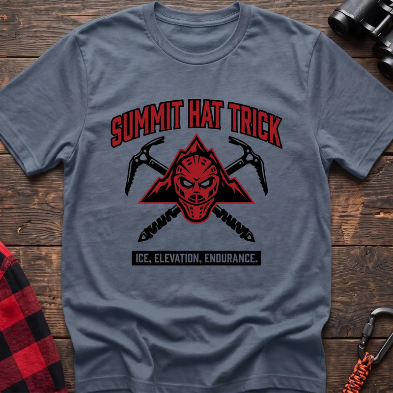 Stone blue t-shirt with a red mountain-themed goalie mask and crossed ice axes, with the text “SUMMIT HAT TRICK” and “ICE, ELEVATION, ENDURANCE,” laid flat on a wooden surface.