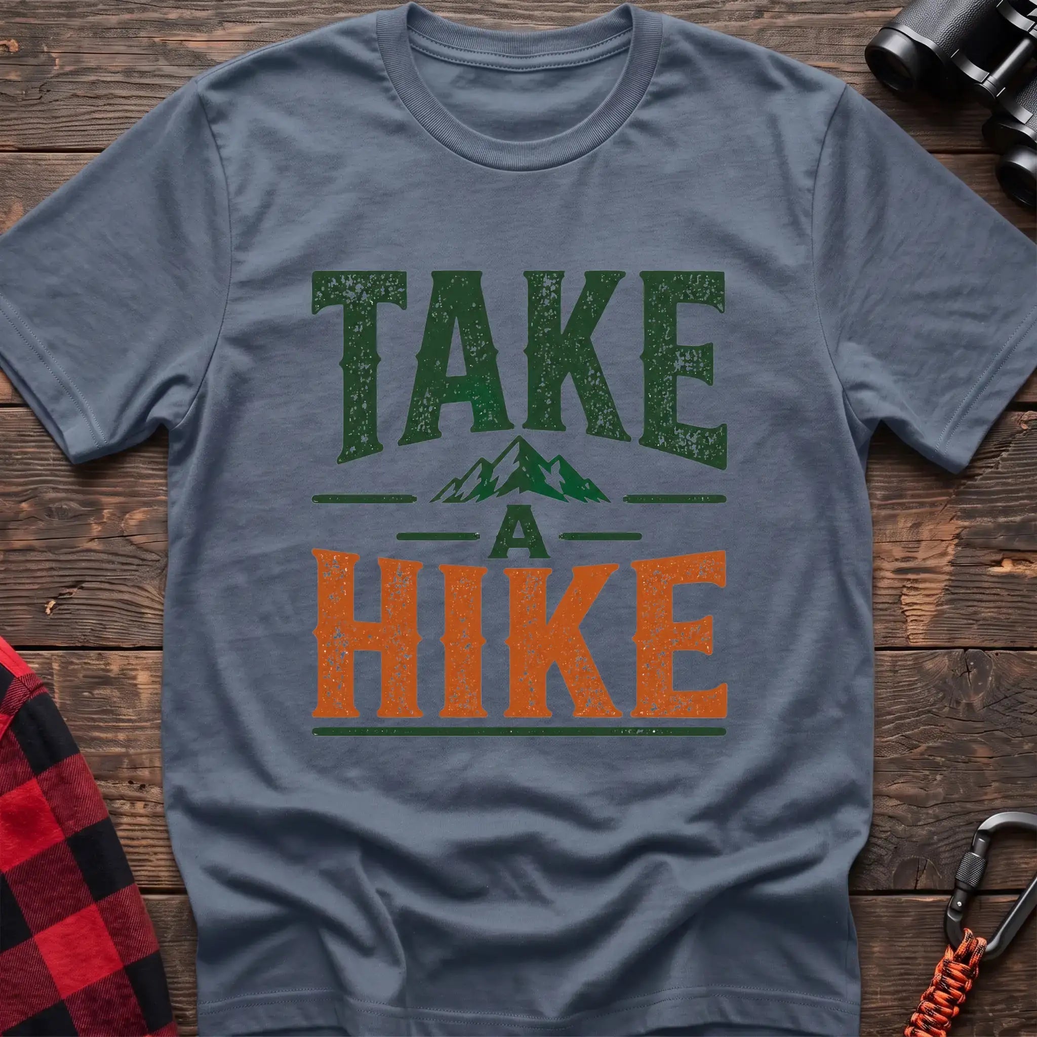 Stone blue t-shirt with distressed green and orange text reading “TAKE A HIKE” with a small mountain graphic between the words, laid flat on a wooden surface.