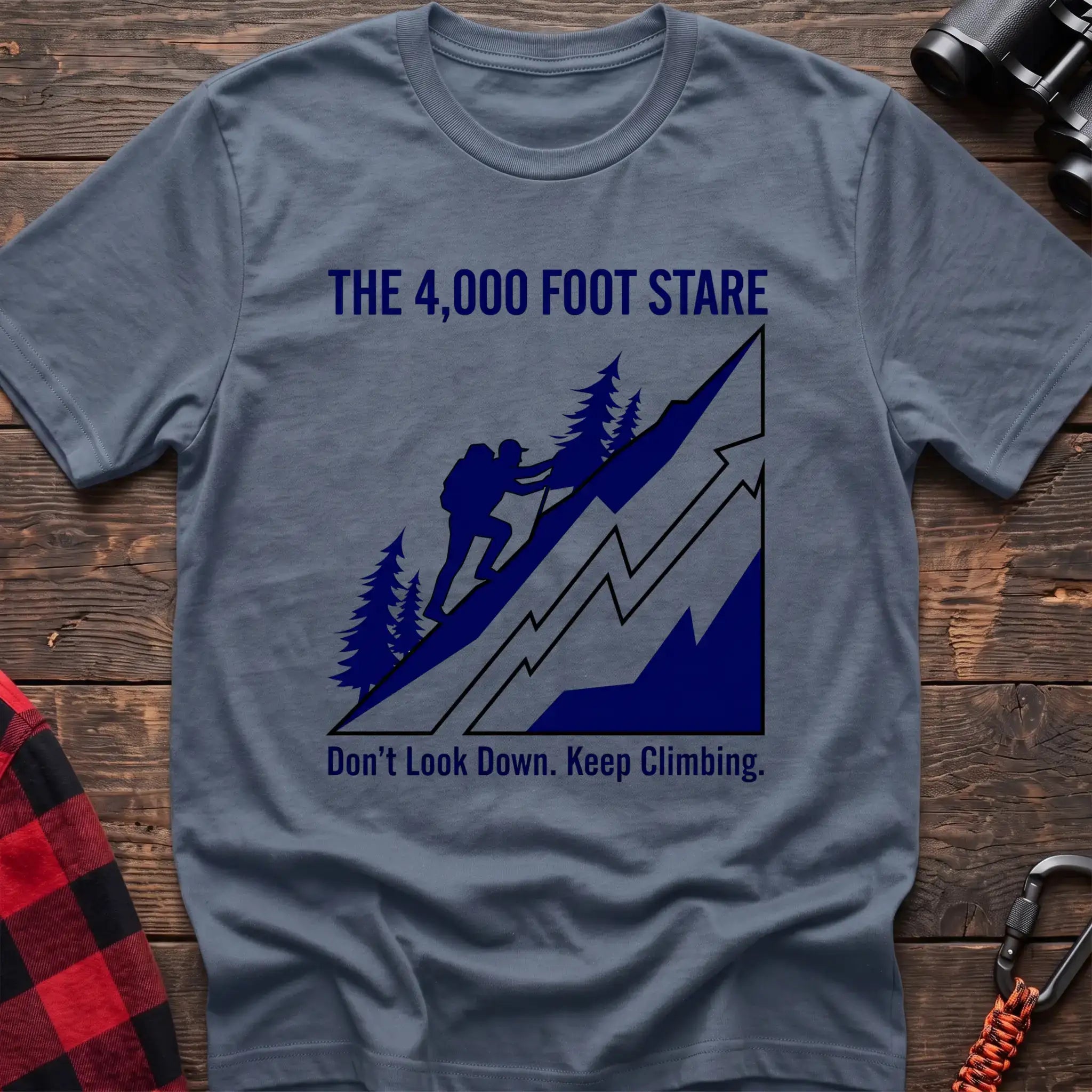 Stone blue t-shirt with a blue illustration of a hiker climbing a steep mountain, with the text “THE 4,000 FOOT STARE” and “Don’t Look Down. Keep Climbing,” laid flat on a wooden surface.