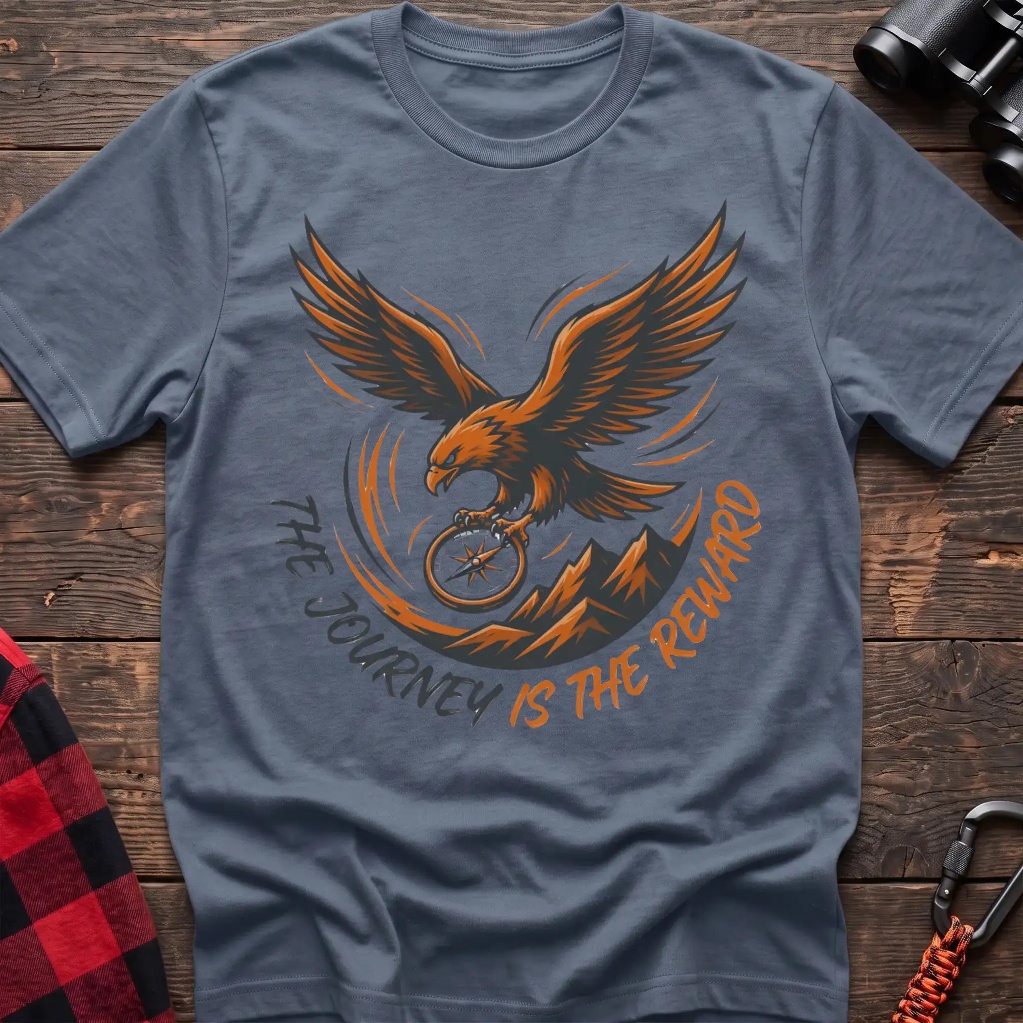 Stone blue t-shirt with an orange and dark graphic of a soaring eagle holding a compass above mountains, with the curved text “THE JOURNEY IS THE REWARD,” laid flat on a wooden surface.