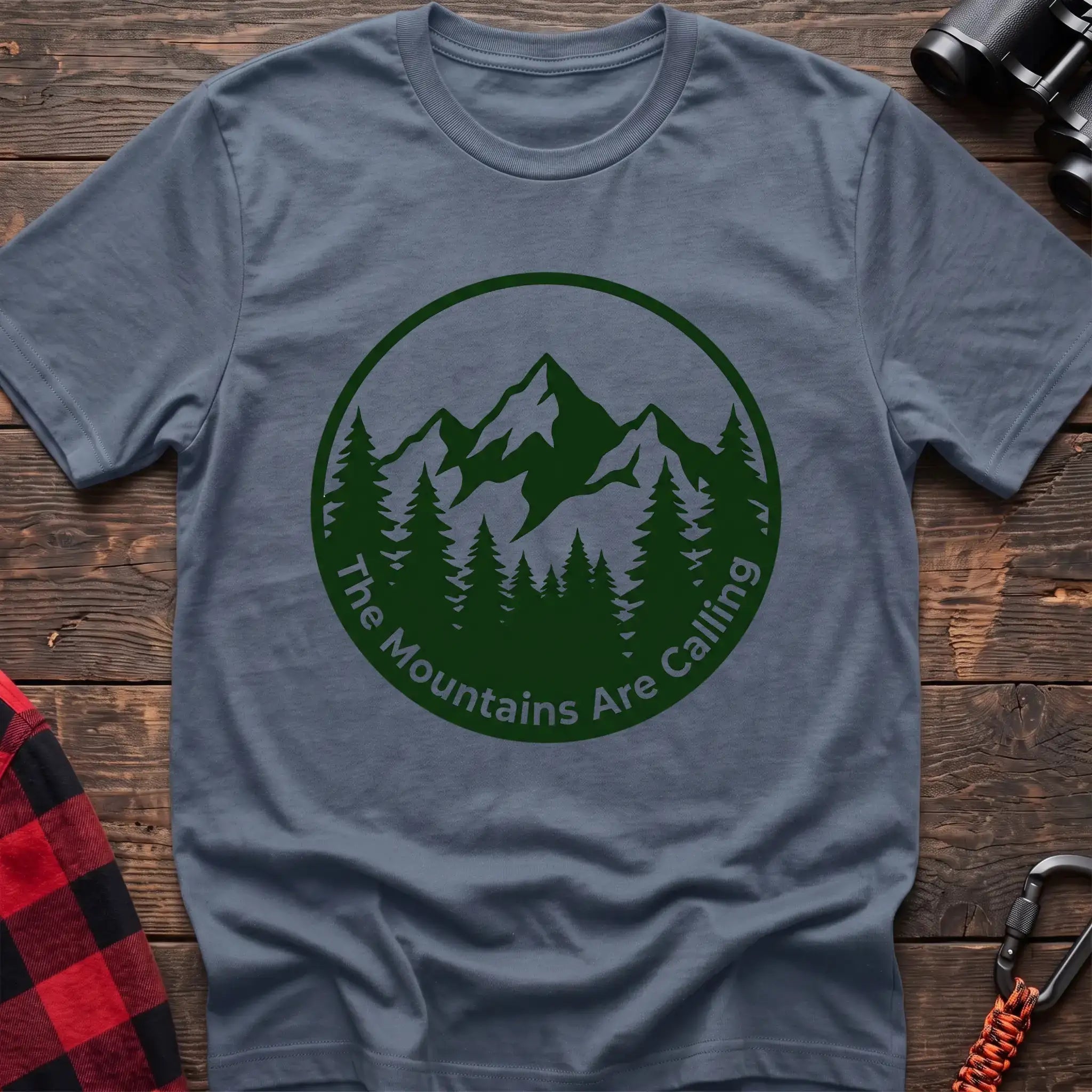 Stone blue t-shirt with a circular dark green design featuring mountain peaks and pine trees, with the text “The Mountains Are Calling,” laid flat on a wooden surface.