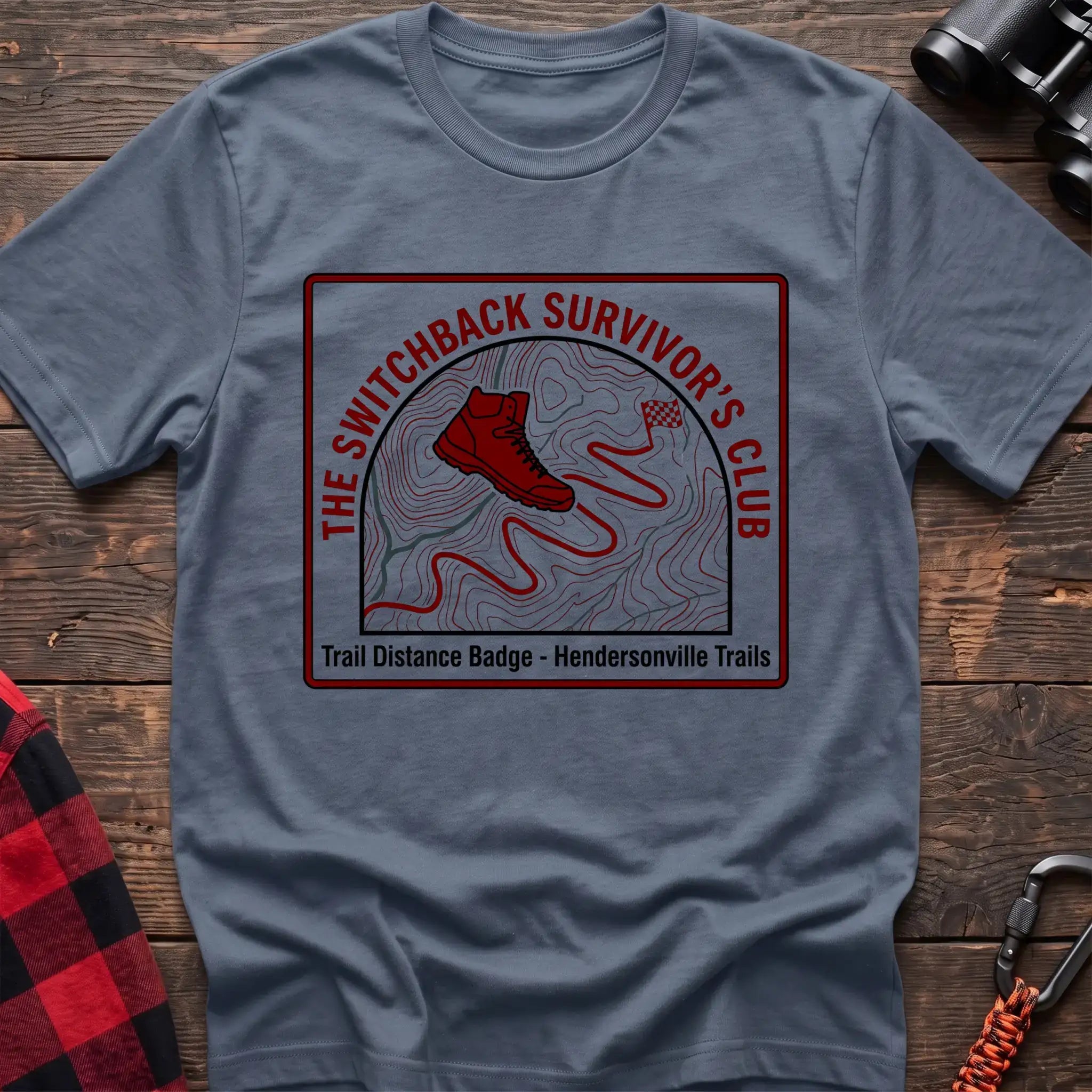 Stone blue t-shirt with a square badge design featuring a red hiking boot on a topographic map with a winding trail, and the text “THE SWITCHBACK SURVIVOR’S CLUB” and “Trail Distance Badge - Hendersonville Trails,” laid flat on a wooden surface.