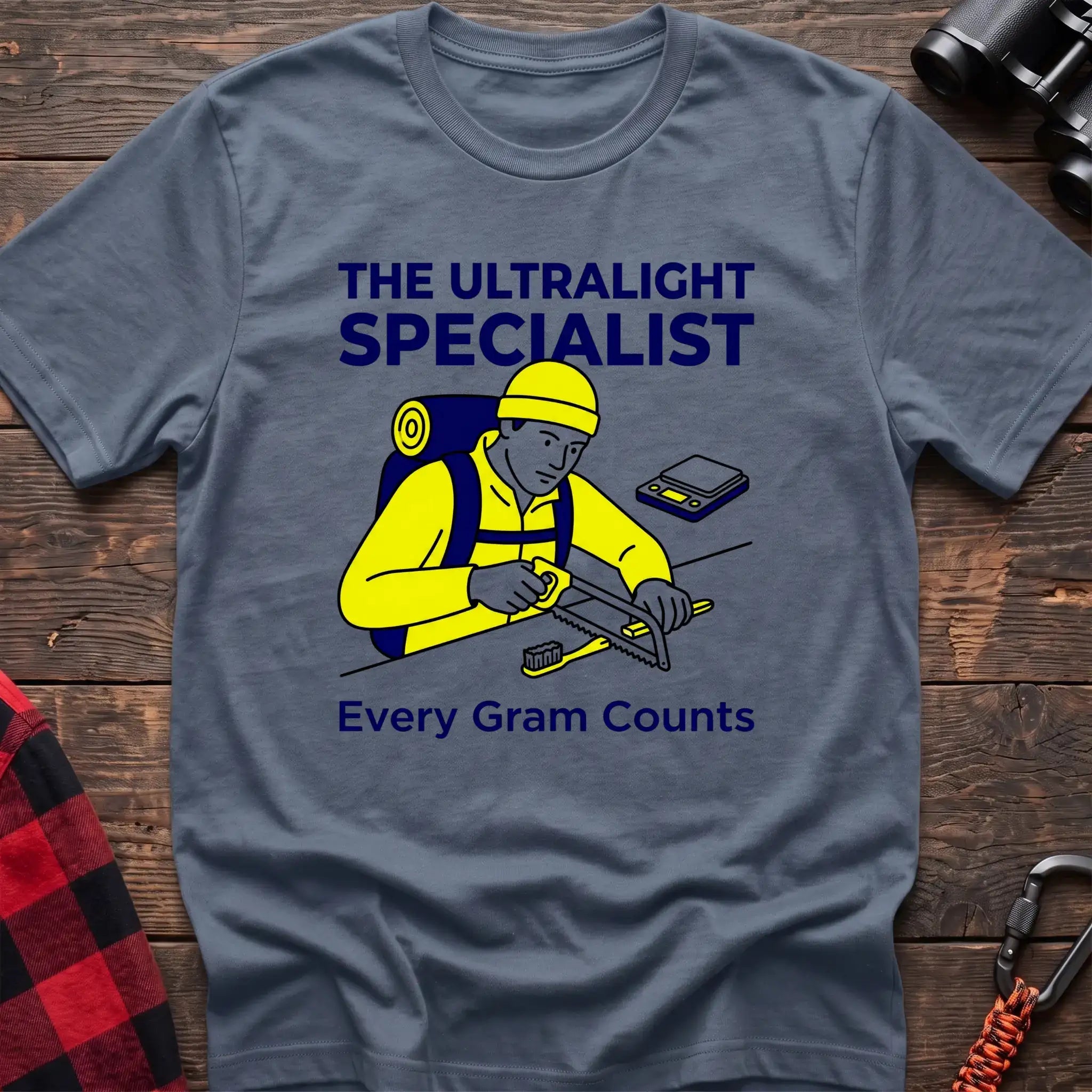 Stone blue t-shirt with an illustration of a hiker sawing a toothbrush on a table next to a scale, with the text “THE ULTRALIGHT SPECIALIST” and “Every Gram Counts,” laid flat on a wooden surface.