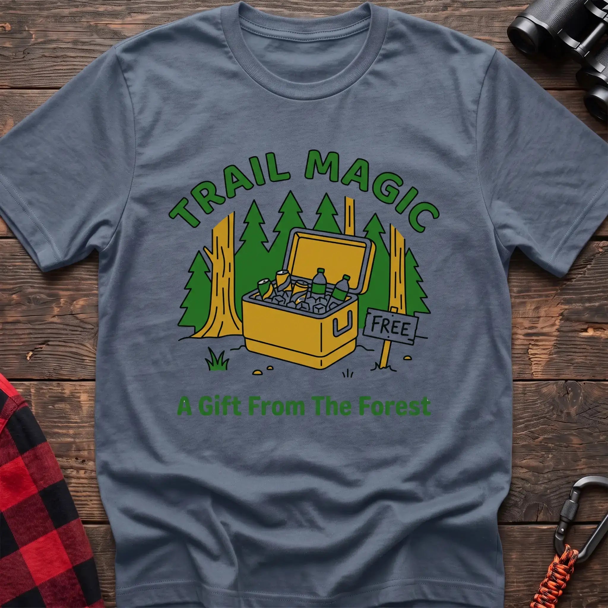 Stone blue t-shirt with an illustration of an open cooler filled with drinks in a forest setting, with the text “TRAIL MAGIC” and “A Gift From The Forest,” laid flat on a wooden surface.