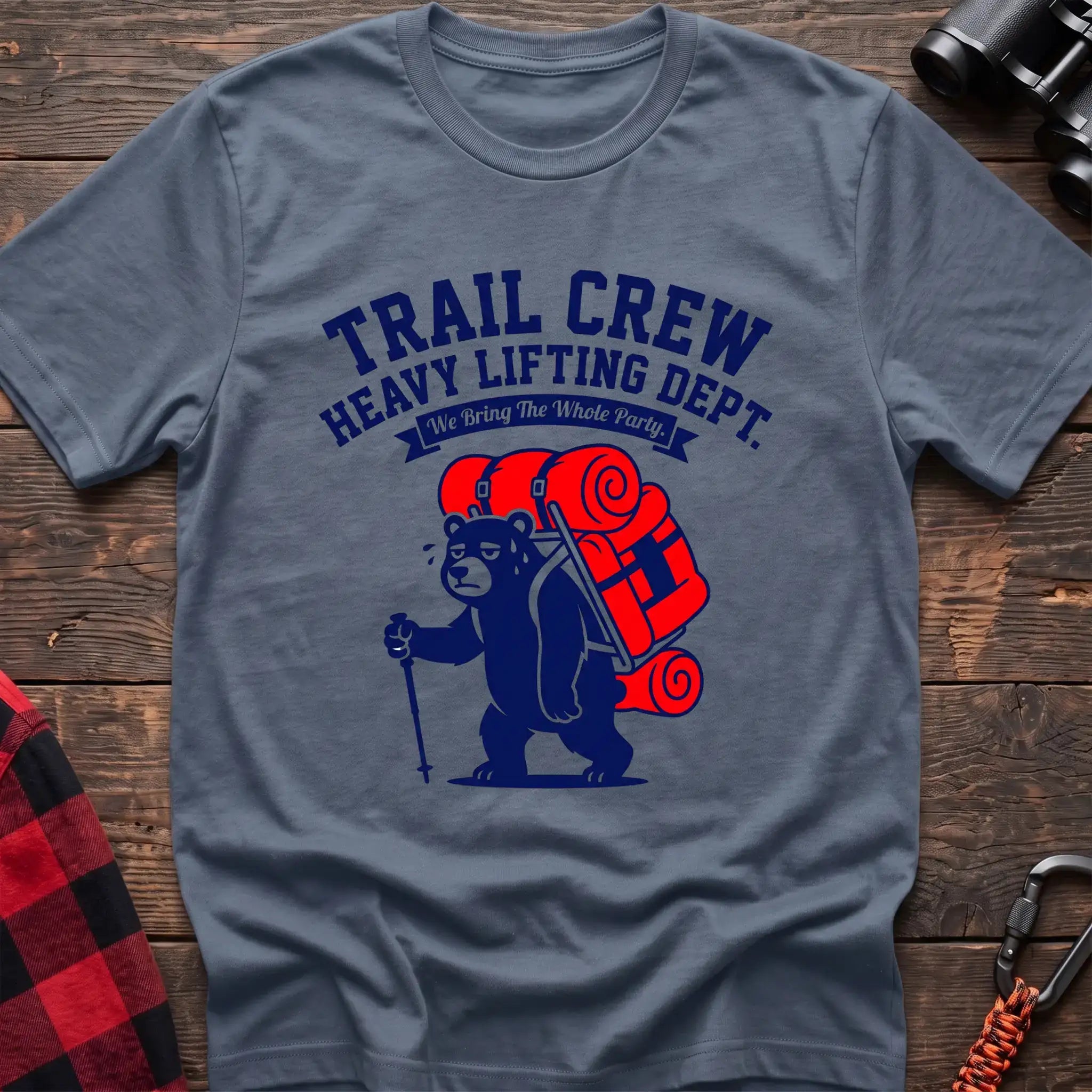 Stone blue t-shirt with a cartoon dark blue bear carrying a large red backpack and hiking pole, with the text “TRAIL CREW HEAVY LIFTING DEPT. We Bring The Whole Party,” laid flat on a wooden surface.