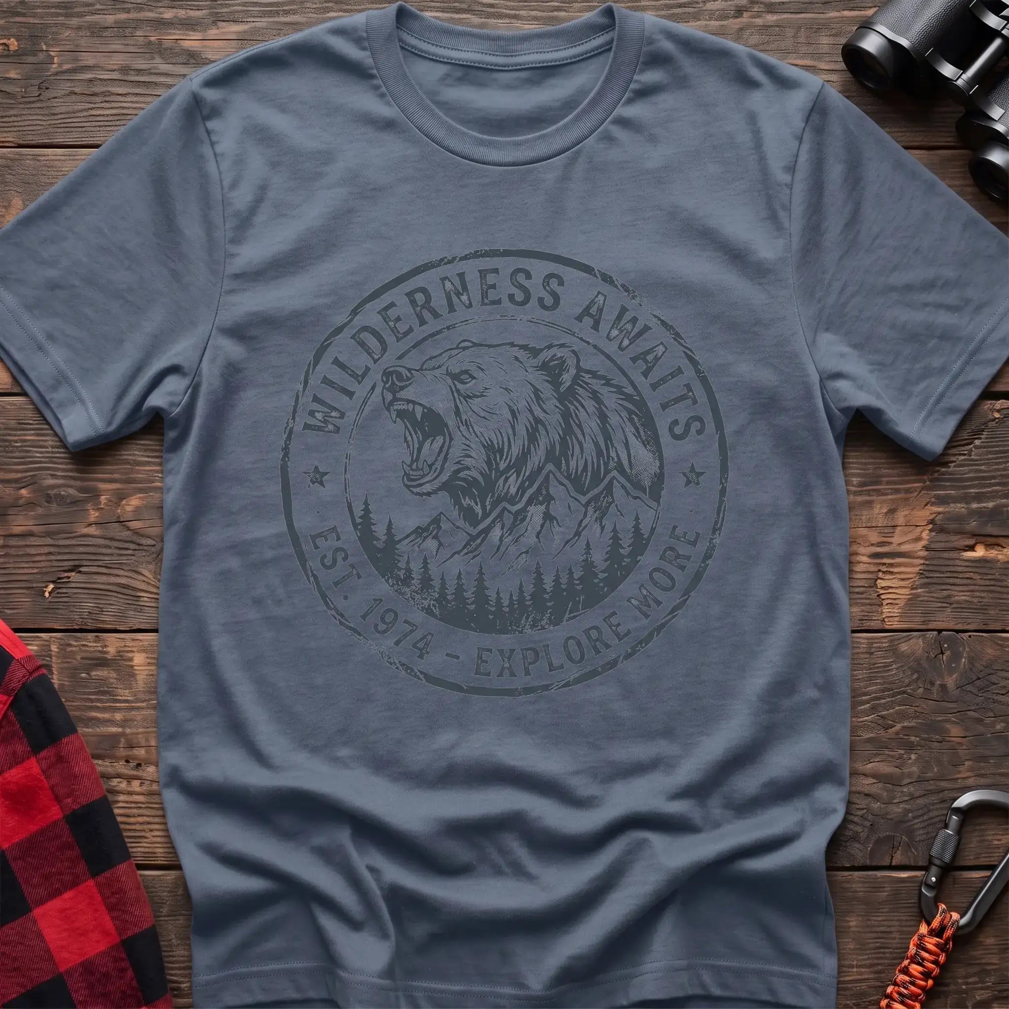 Stone blue t-shirt with a circular dark gray badge design featuring a roaring bear head above mountains and pine trees, with the text “WILDERNESS AWAITS” and “EST. 1974 - EXPLORE MORE,” laid flat on a wooden surface.