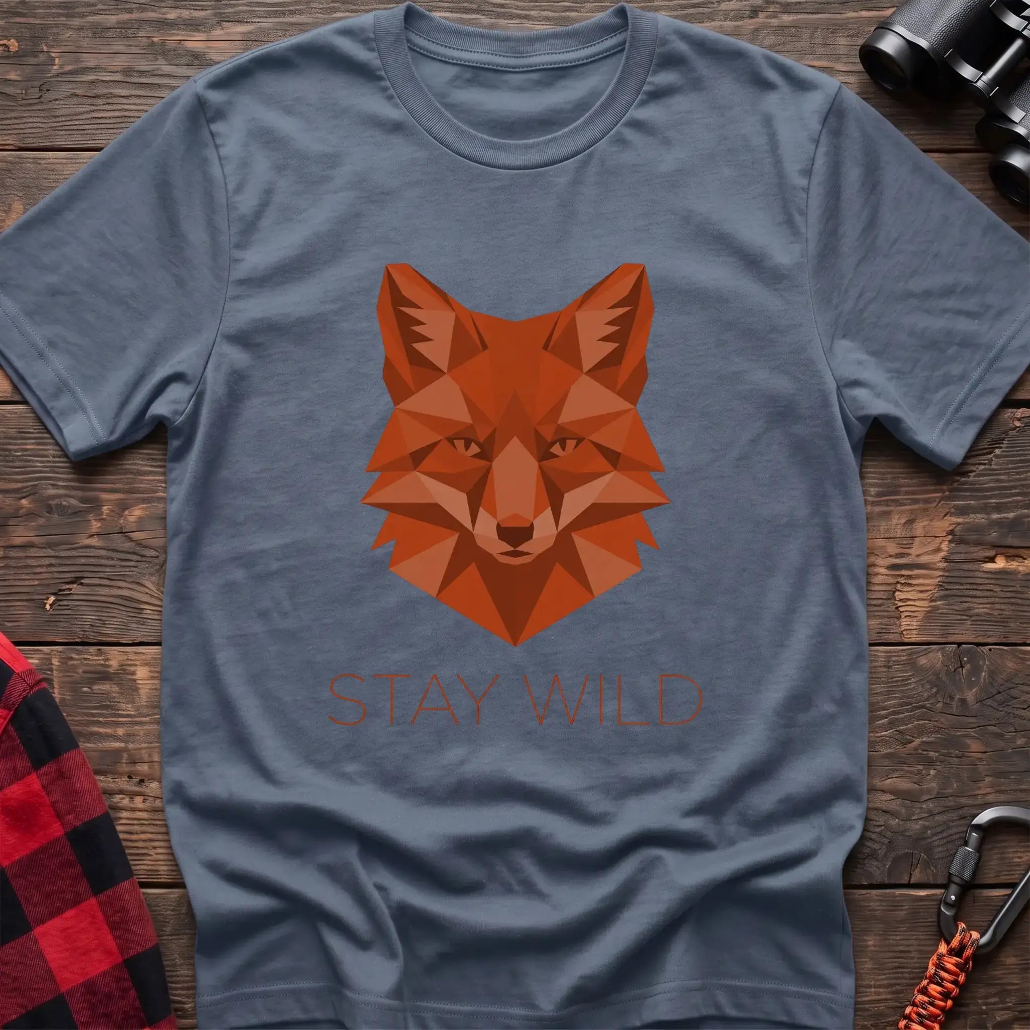 Stone blue t-shirt with a geometric low-poly orange fox head and the text “STAY WILD” printed below in matching orange, laid flat on a wooden surface.