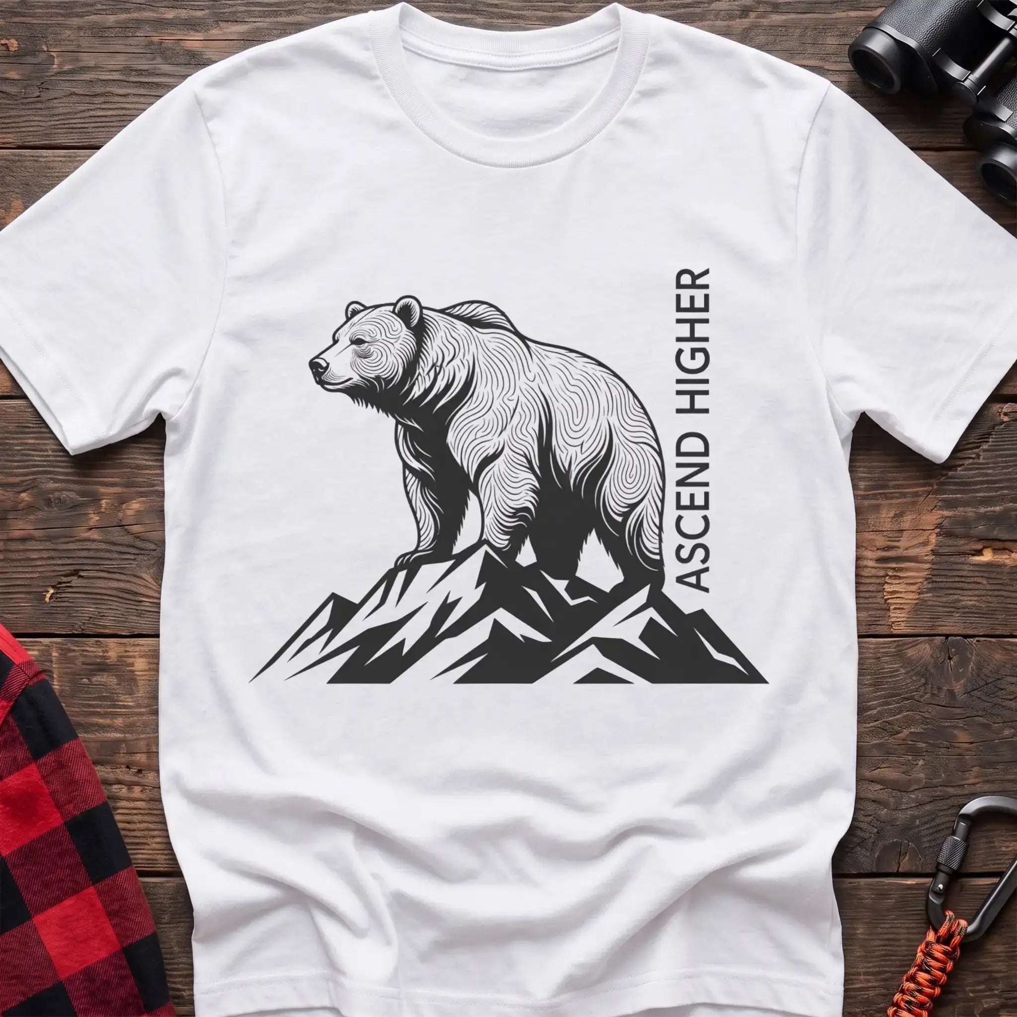 White t-shirt with a black illustration of a bear standing on mountain peaks, with the vertical text “ASCEND HIGHER” beside it, laid flat on a wooden surface.