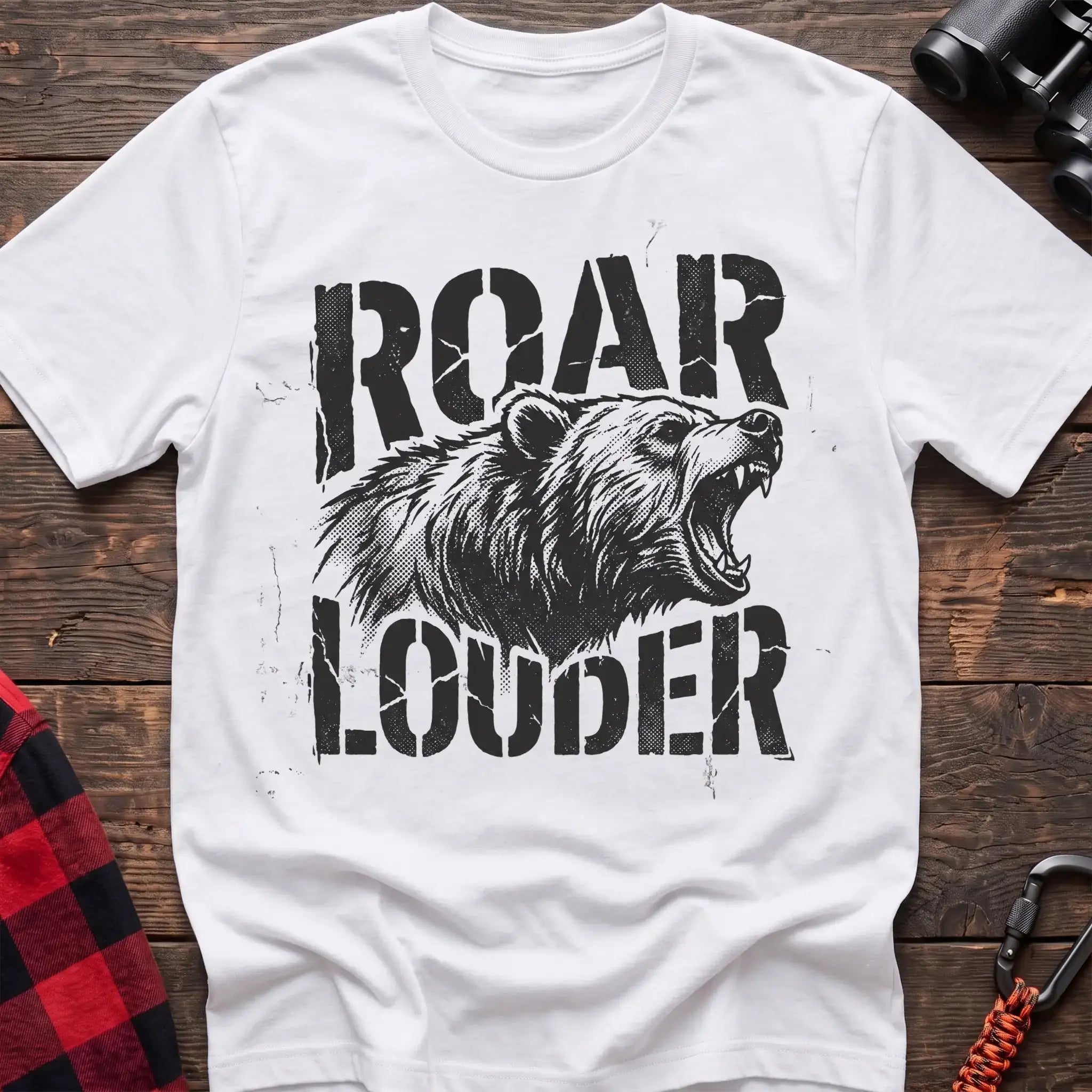 White t-shirt with a distressed black graphic of a roaring bear head between the bold text “ROAR” and “LOUDER,” laid flat on a wooden surface.