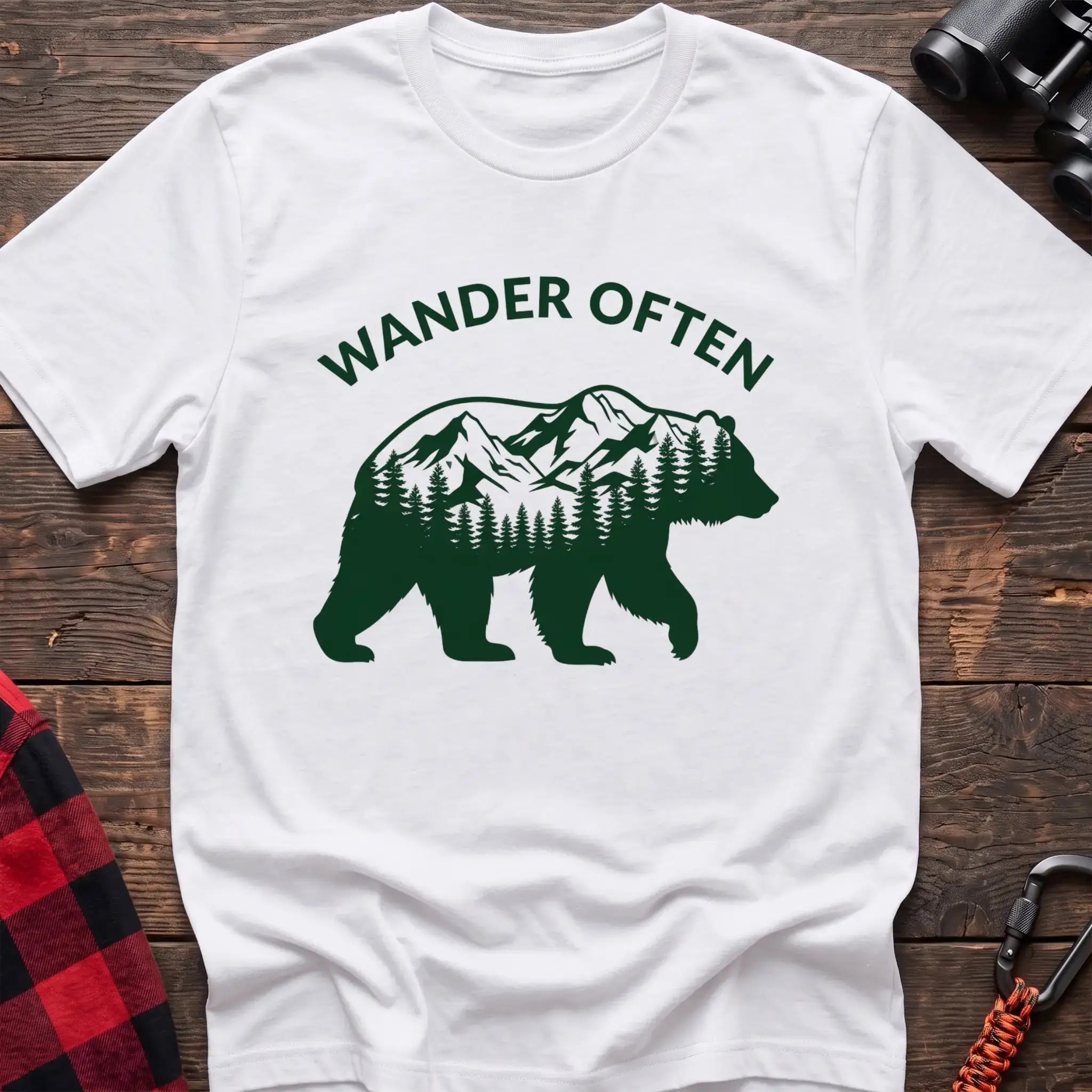White t-shirt with a dark green silhouette of a walking bear containing a mountain and pine forest scene inside, with the arched text “WANDER OFTEN” above it, laid flat on a wooden surface.