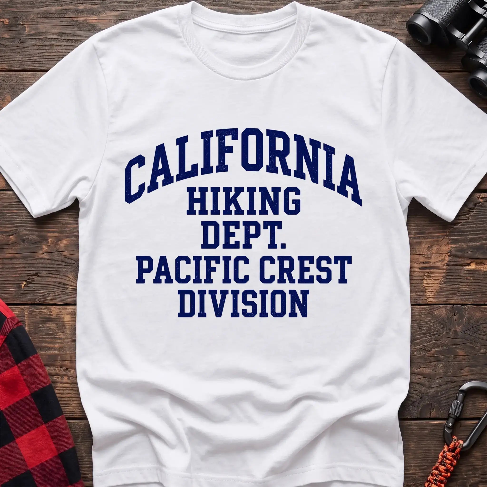 White t-shirt with distressed blue collegiate-style text reading “CALIFORNIA HIKING DEPT. PACIFIC CREST DIVISION,” laid flat on a wooden surface.