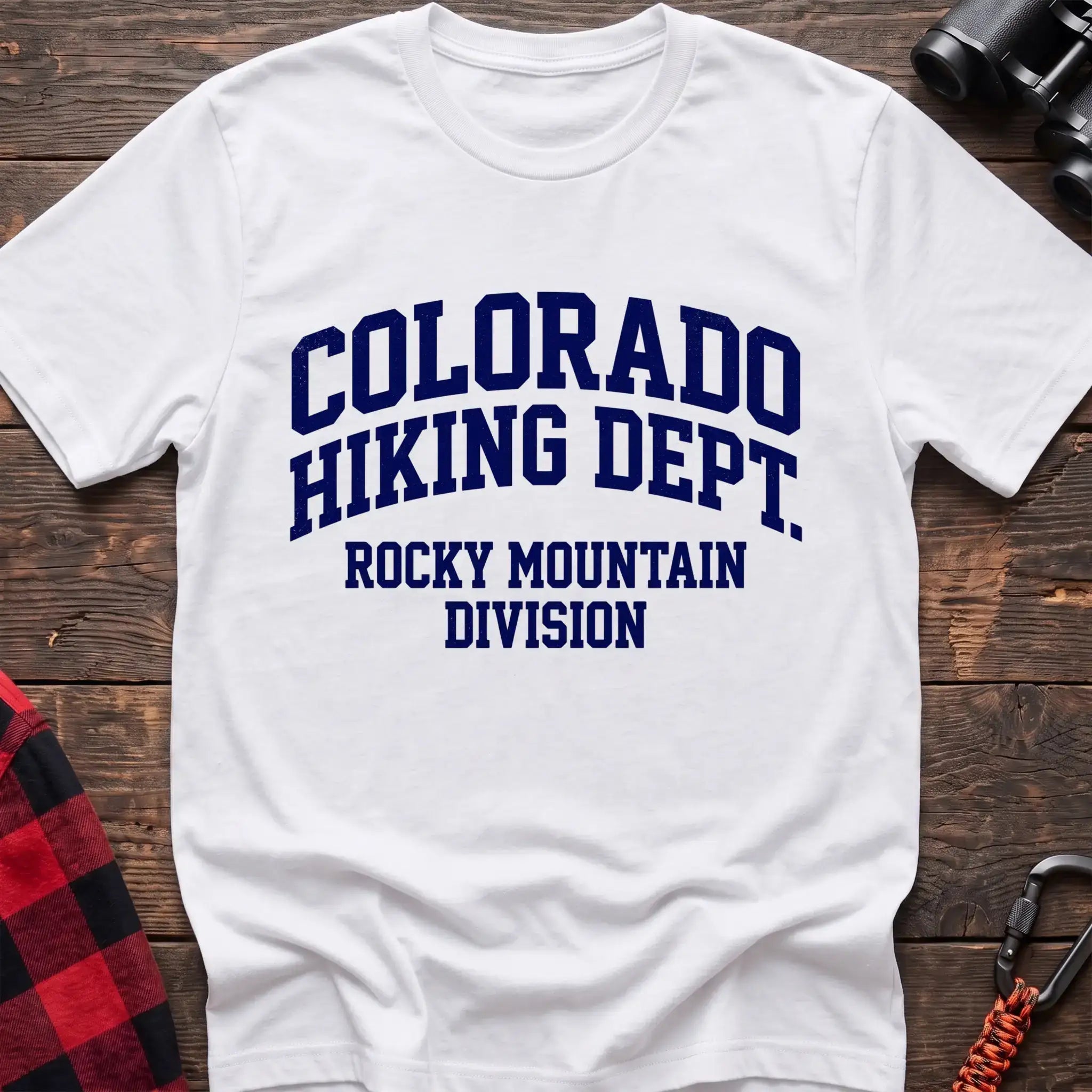 White t-shirt with distressed dark blue collegiate-style text reading “COLORADO HIKING DEPT. ROCKY MOUNTAIN DIVISION,” laid flat on a wooden surface.