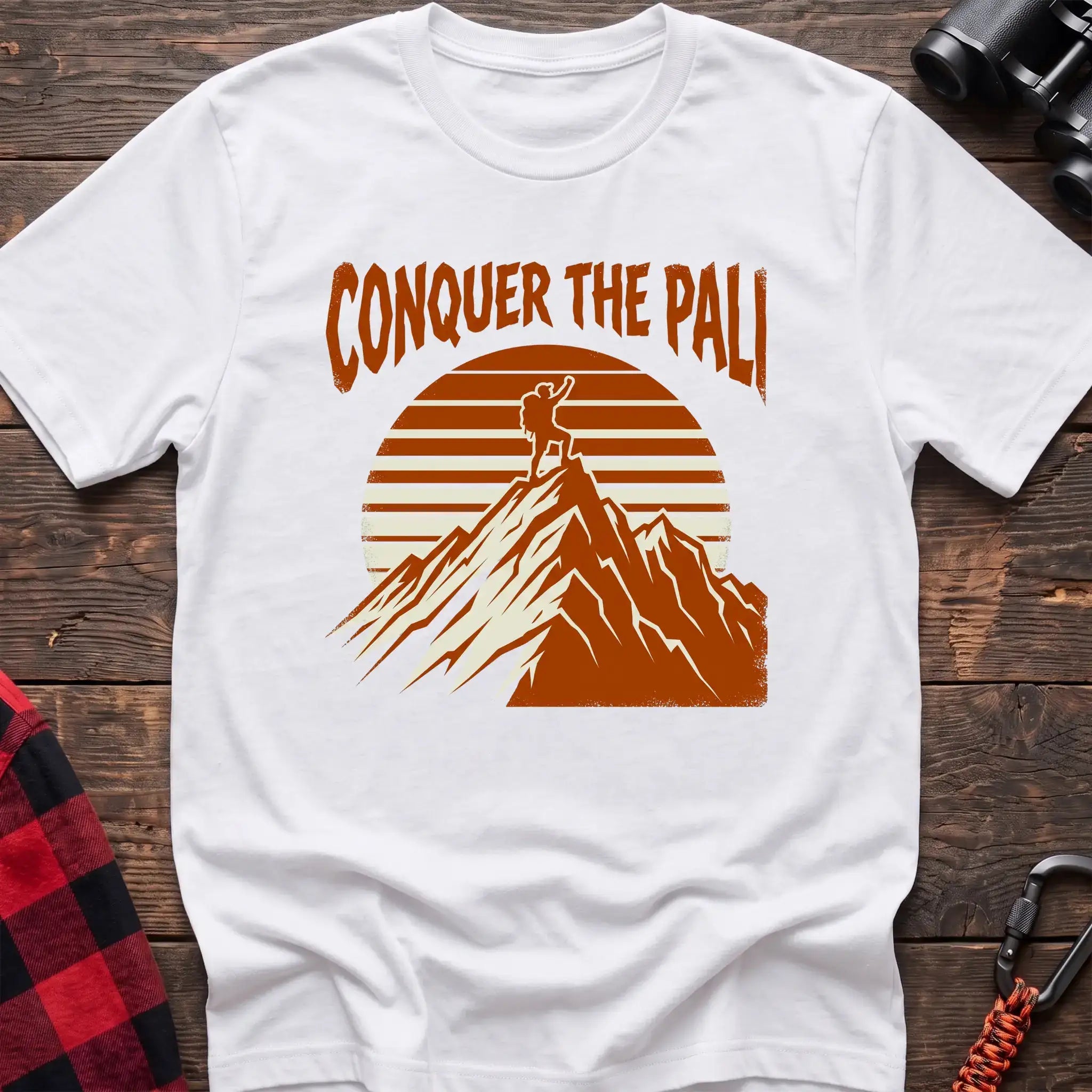 White t-shirt with orange text reading Conquer the Pali above a hiker standing on a mountain peak with a retro sun graphic