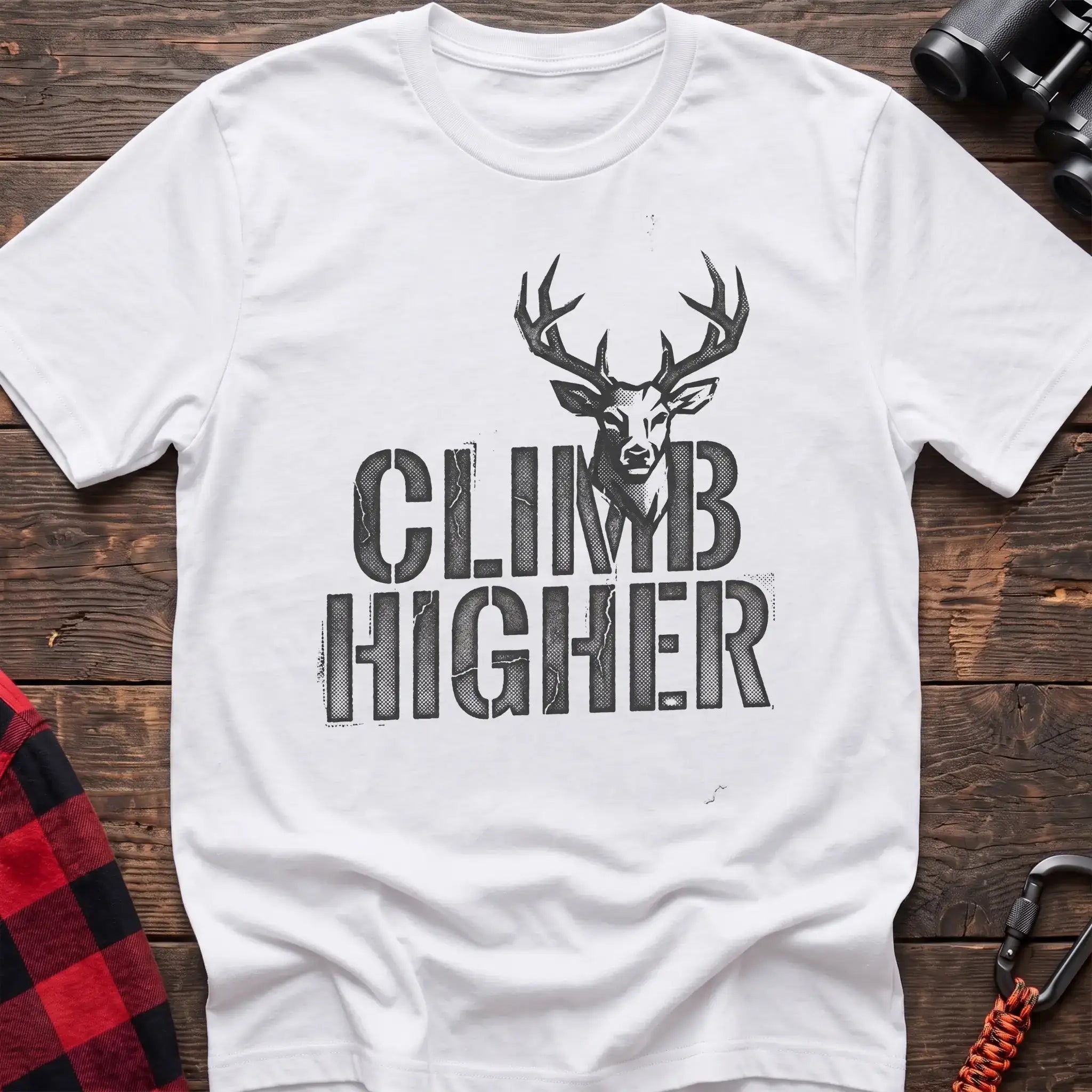 White t-shirt with a distressed dark graphic of a deer’s head and antlers rising from the bold text “CLIMB HIGHER,” laid flat on a wooden surface.