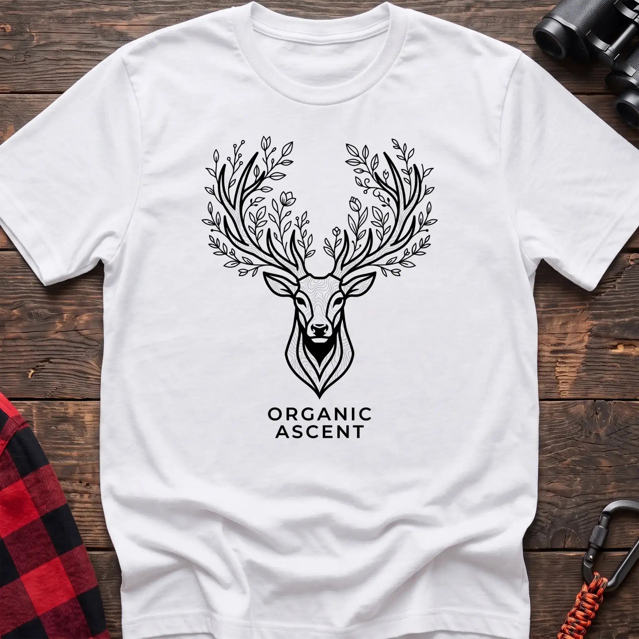 White t-shirt with a black line drawing of a deer’s head whose antlers are formed from leafy branches and flowers, with the text “ORGANIC ASCENT” printed below, laid flat on a wooden surface.