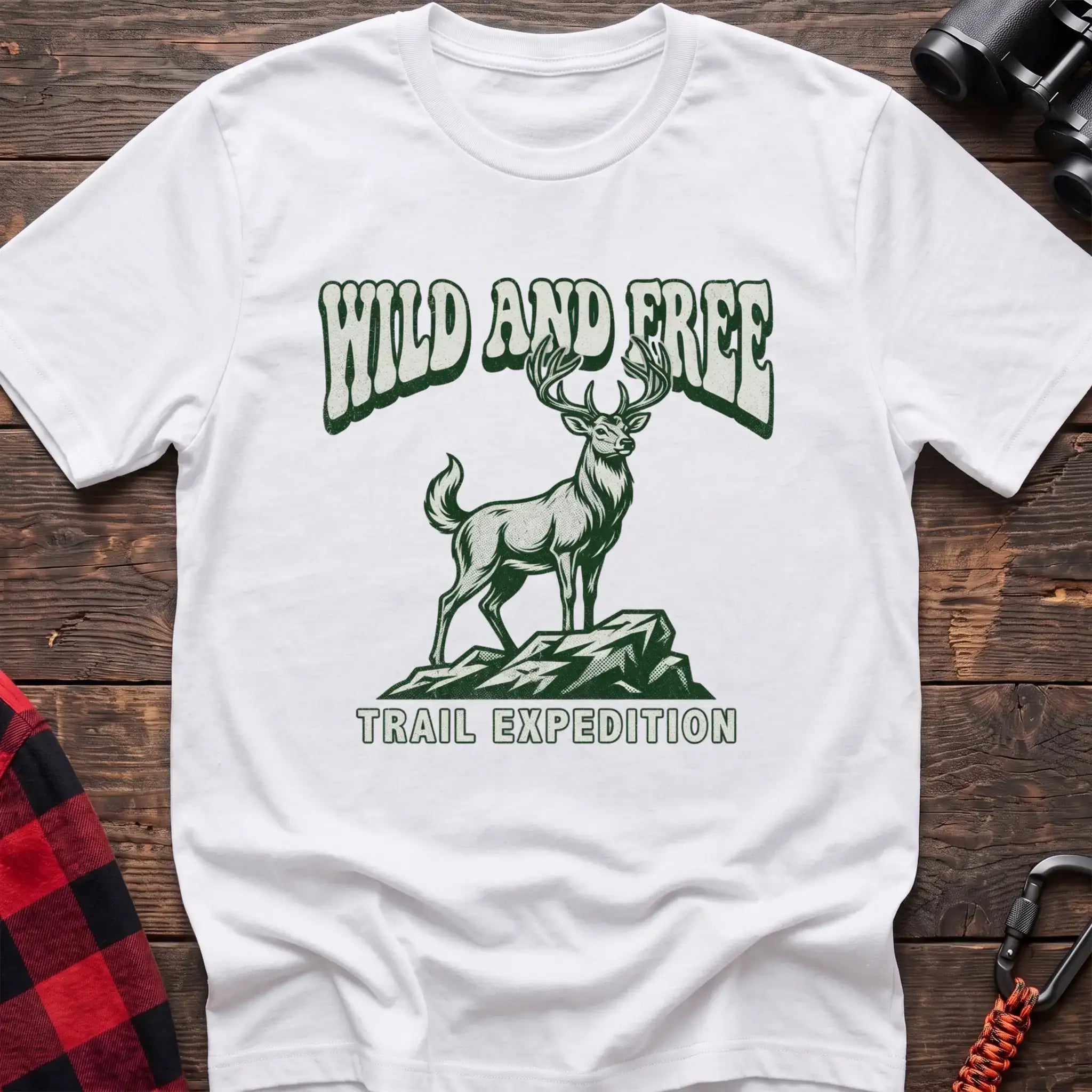 White t-shirt with a rustic graphic of a deer standing on a rocky outcrop, with the arched text “WILD AND FREE” above and “TRAIL EXPEDITION” below, laid flat on a wooden surface.