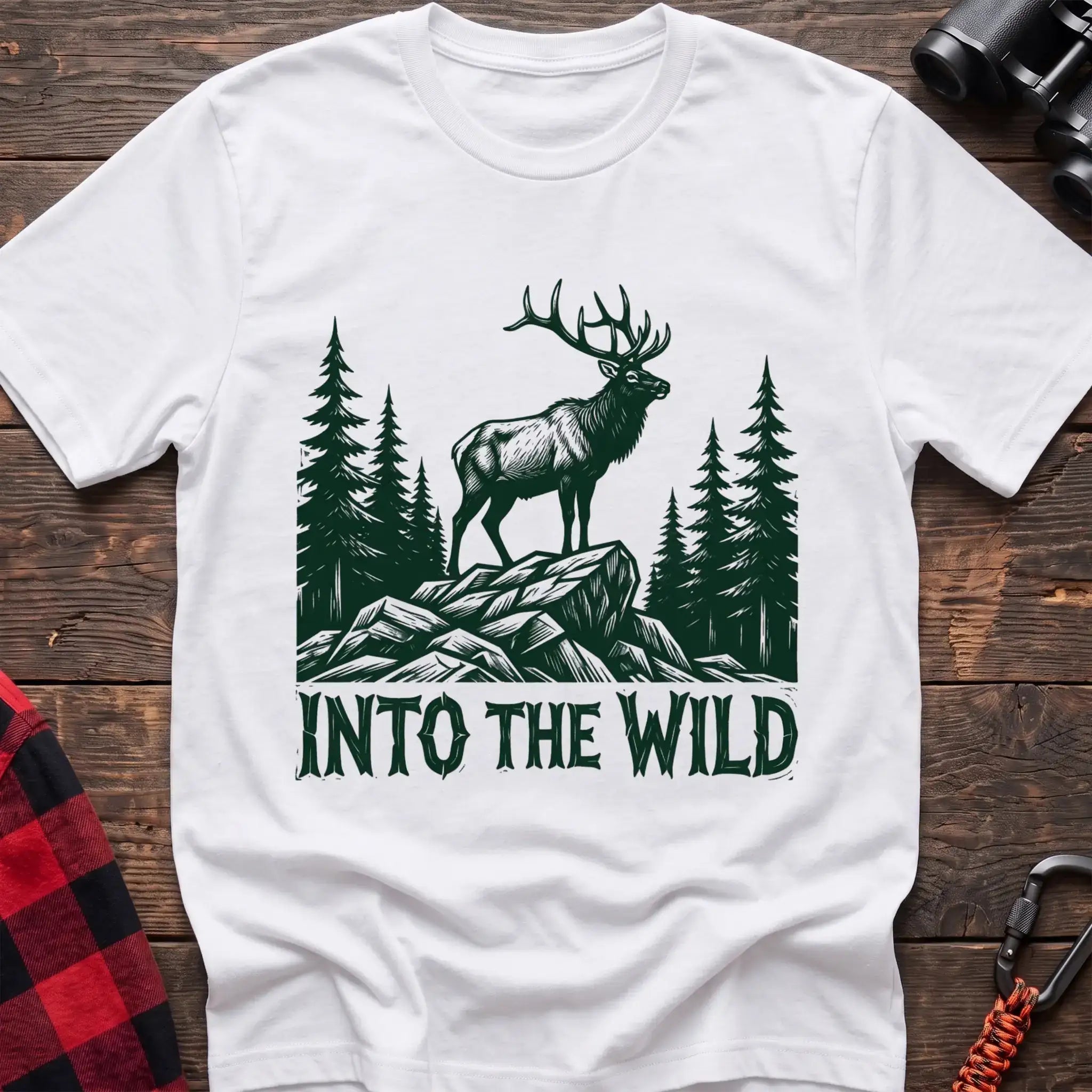 White t-shirt with a dark green illustration of an elk standing on a rocky outcrop among pine trees, with the text “INTO THE WILD” printed below, laid flat on a wooden surface.