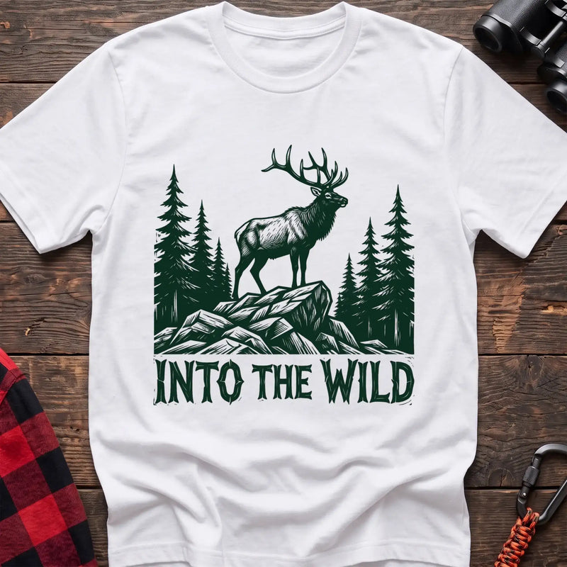 White t-shirt with a dark green illustration of an elk standing on a rocky outcrop among pine trees, with the text “INTO THE WILD” printed below, laid flat on a wooden surface.