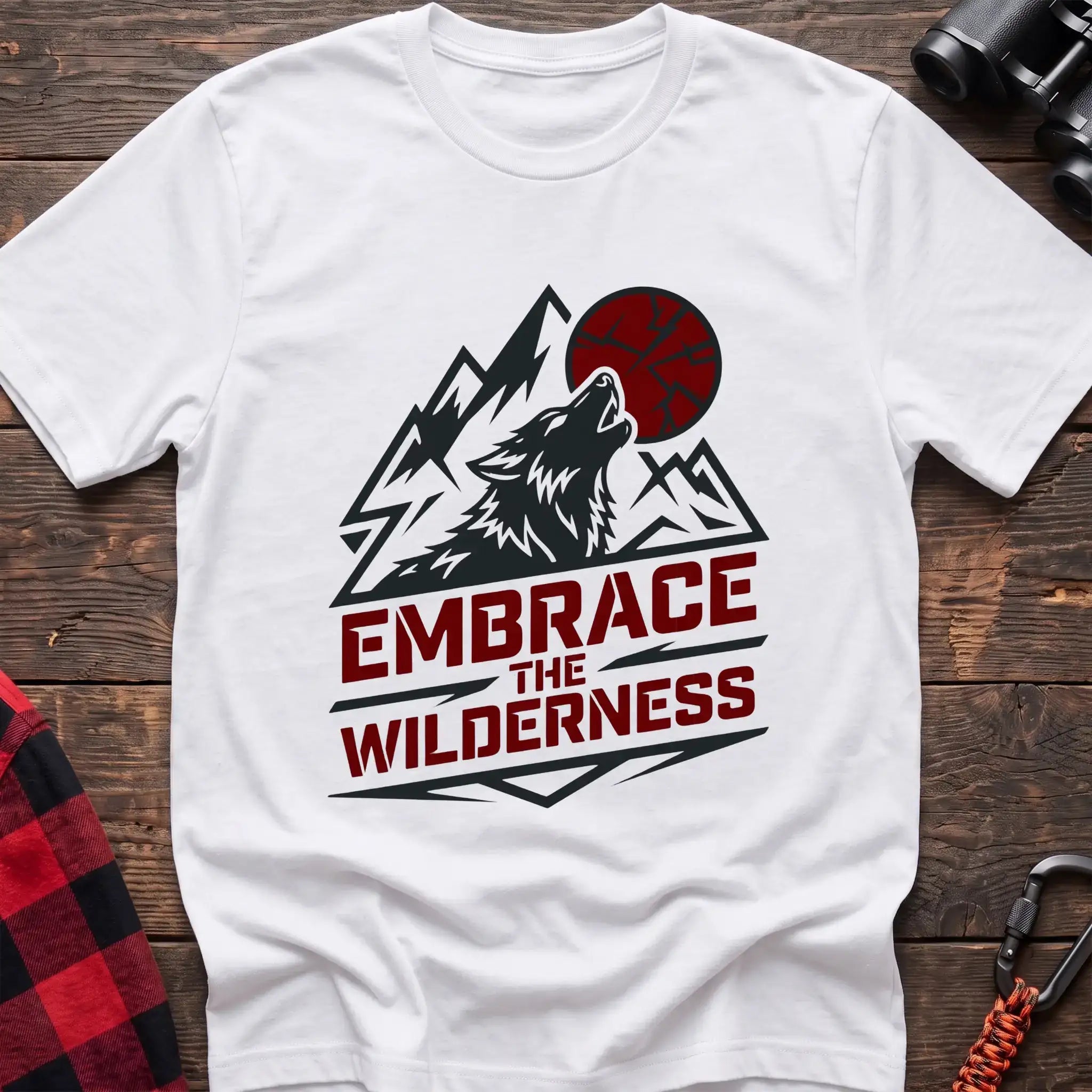 White t-shirt with a dark graphic of a howling wolf against mountain peaks and a red sun, with the bold red text “EMBRACE THE WILDERNESS” below, laid flat on a wooden surface.
