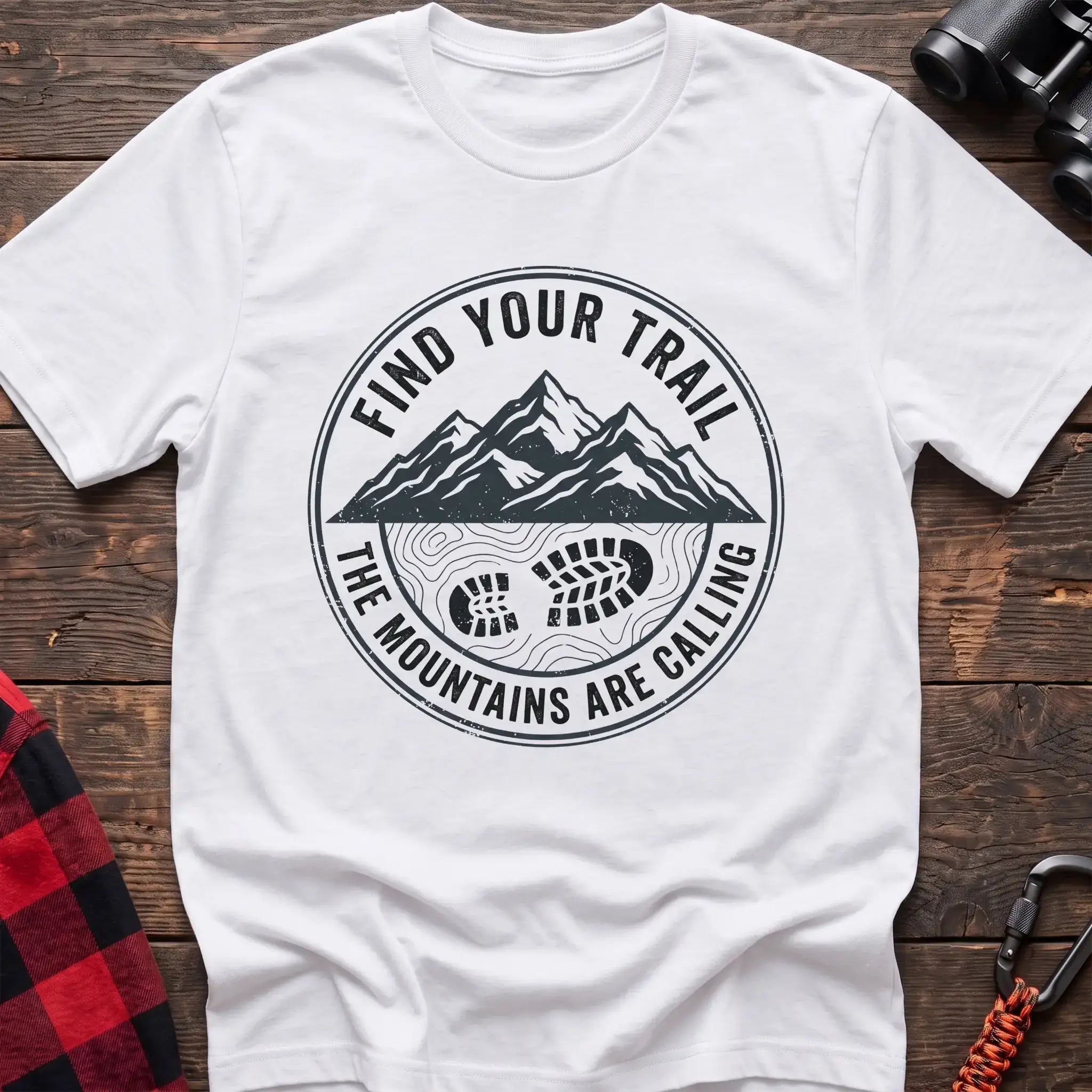 White t-shirt with a circular dark gray badge design featuring mountain peaks above a hiking boot print on topographic lines, with the text “FIND YOUR TRAIL” and “THE MOUNTAINS ARE CALLING,” laid flat on a wooden surface.