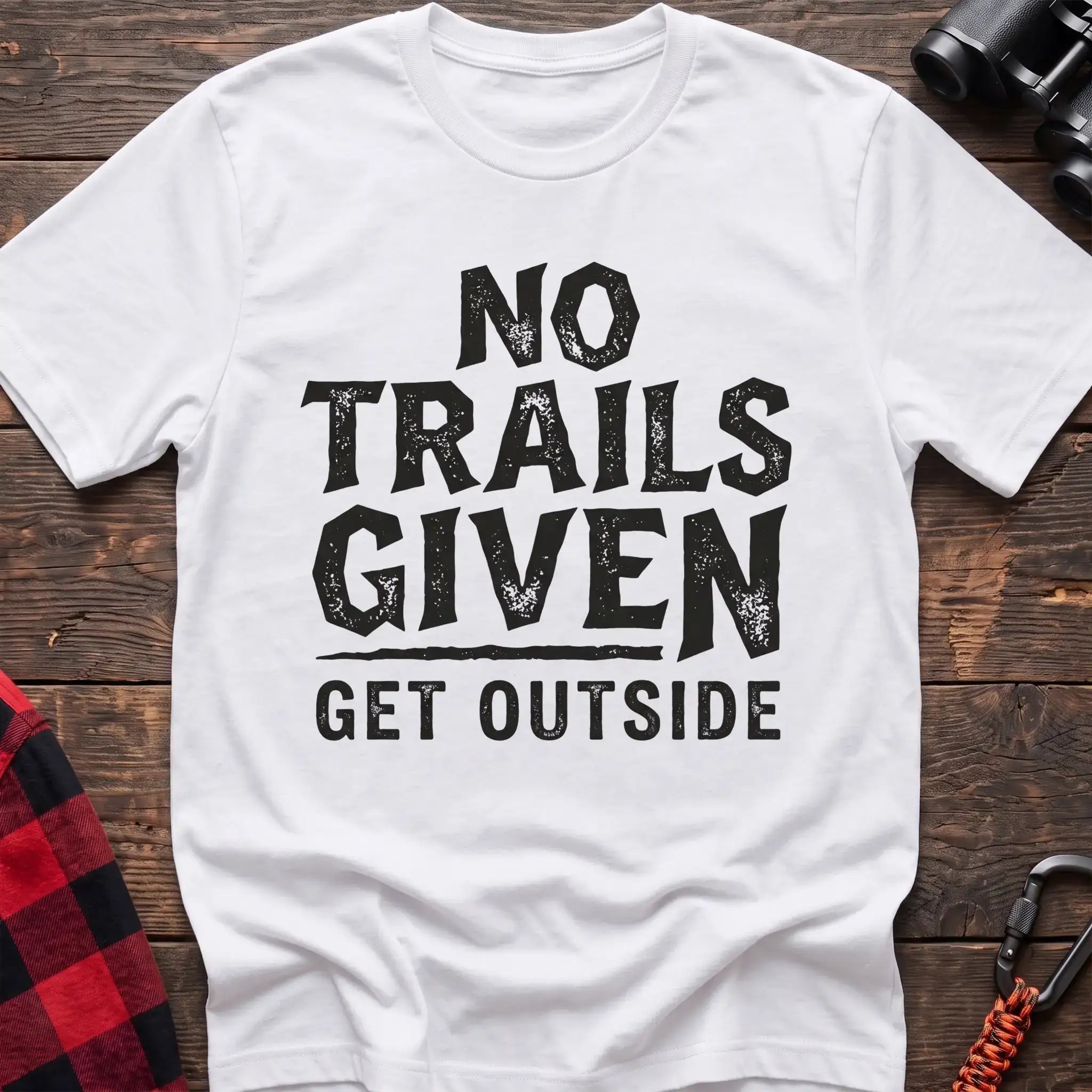 White t-shirt with distressed black text reading “NO TRAILS GIVEN” and “GET OUTSIDE” below it, laid flat on a wooden surface.