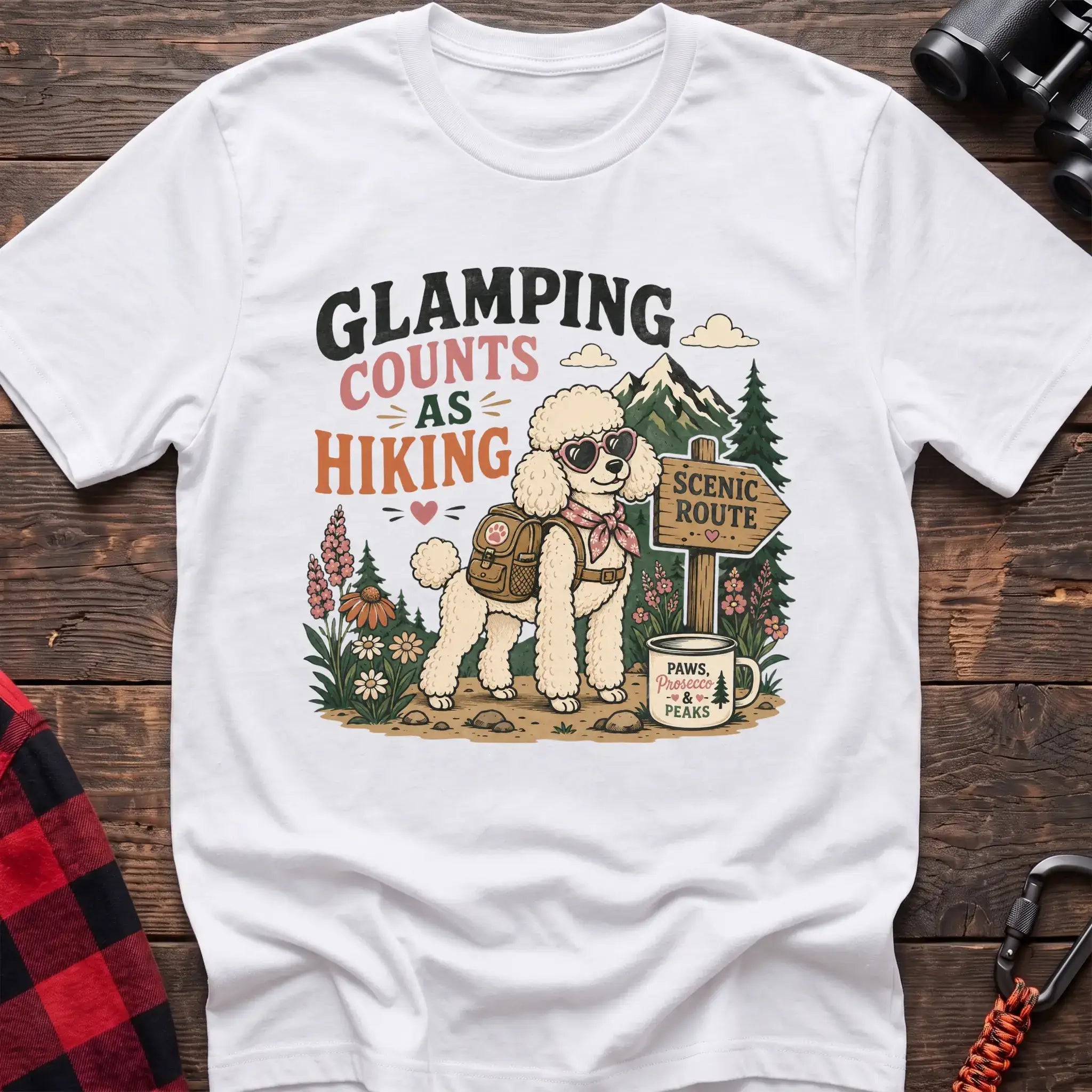 White t-shirt with a colorful illustration of a poodle in sunglasses and a backpack on a scenic trail, with the text “GLAMPING COUNTS AS HIKING,” laid flat on a wooden surface.