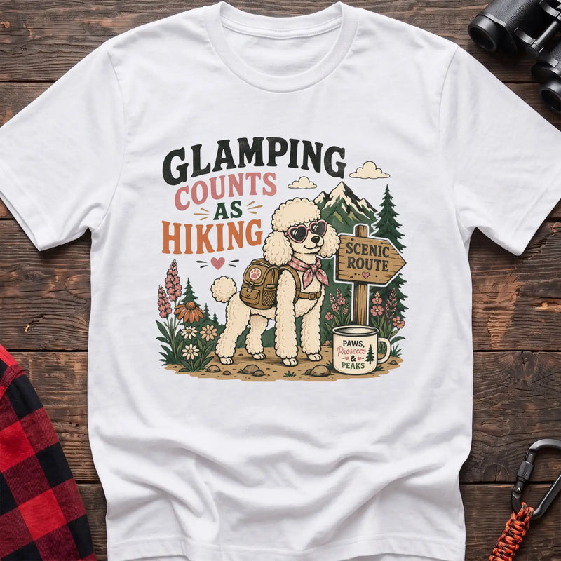 White t-shirt with a colorful illustration of a poodle in sunglasses and a backpack on a scenic trail, with the text “GLAMPING COUNTS AS HIKING,” laid flat on a wooden surface.