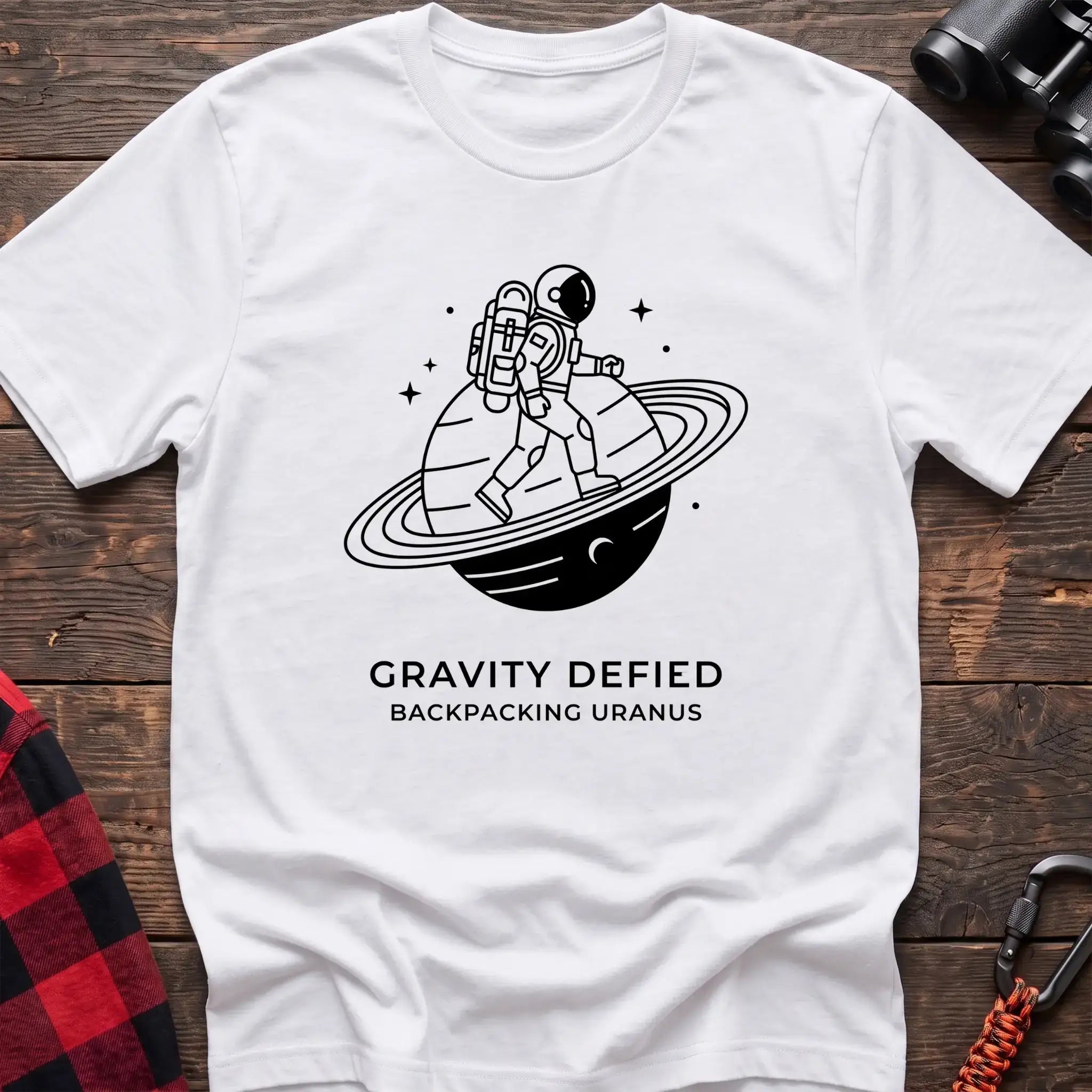 White crew-neck t-shirt with black line-art graphic of an astronaut backpacking across a ringed planet surrounded by stars, with the text “GRAVITY DEFIED / BACKPACKING URANUS” printed below, laid flat on a wooden surface.