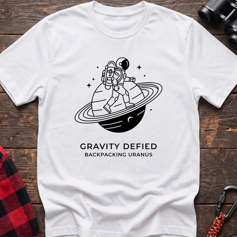 White crew-neck t-shirt with black line-art graphic of an astronaut backpacking across a ringed planet surrounded by stars, with the text “GRAVITY DEFIED / BACKPACKING URANUS” printed below, laid flat on a wooden surface.