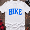 White cotton t-shirt with distressed arched blue HIKE graphic in cracked varsity lettering, flat lay on rustic wood with hiking gear