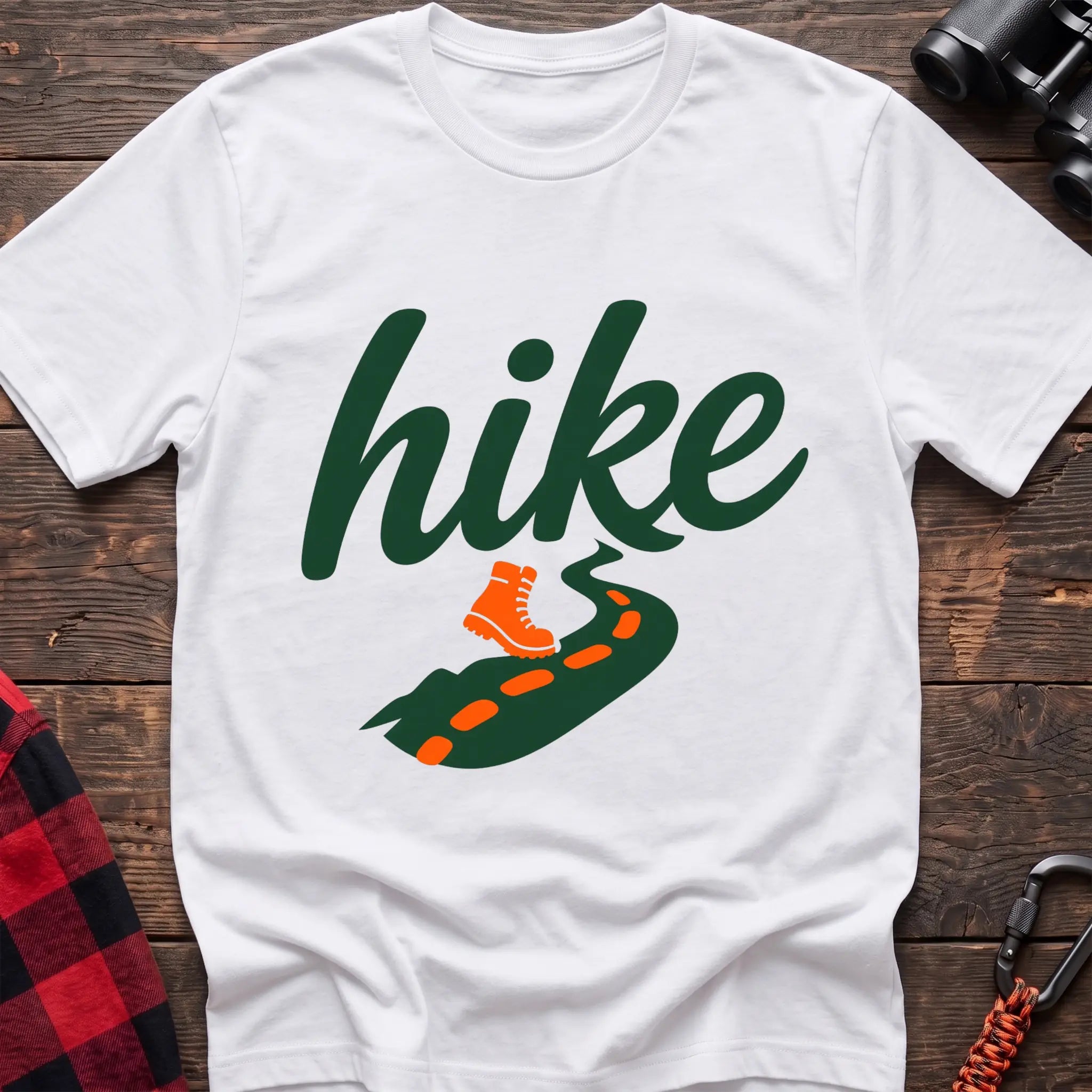 White cotton t-shirt with green cursive hike script, orange hiking boot, and footprint trail graphic, flat lay on rustic wood with hiking gear