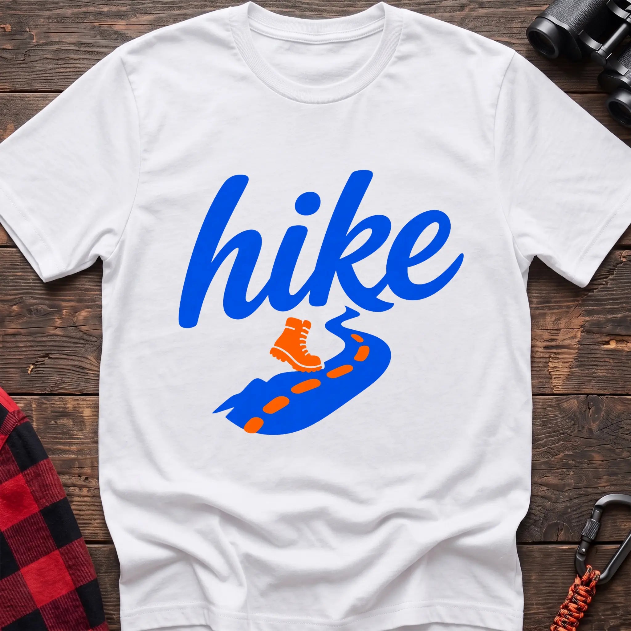 White cotton t-shirt with royal blue cursive hike script, orange hiking boot, and blue trail with orange footprints, flat lay on rustic wood with hiking gear