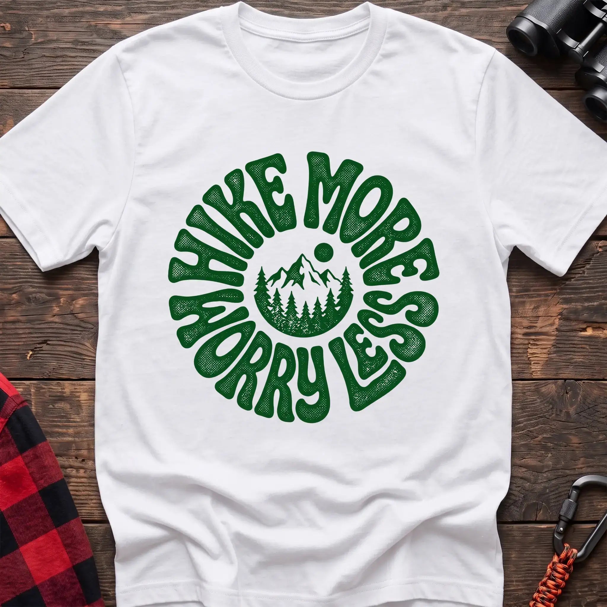 White t-shirt with a circular green design featuring mountains and pine trees in the center, surrounded by the text “HIKE MORE WORRY LESS,” laid flat on a wooden surface.