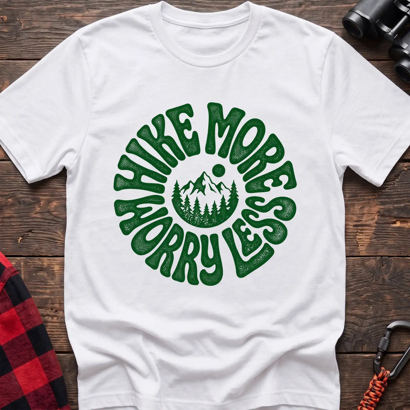 White t-shirt with a circular green design featuring mountains and pine trees in the center, surrounded by the text “HIKE MORE WORRY LESS,” laid flat on a wooden surface.