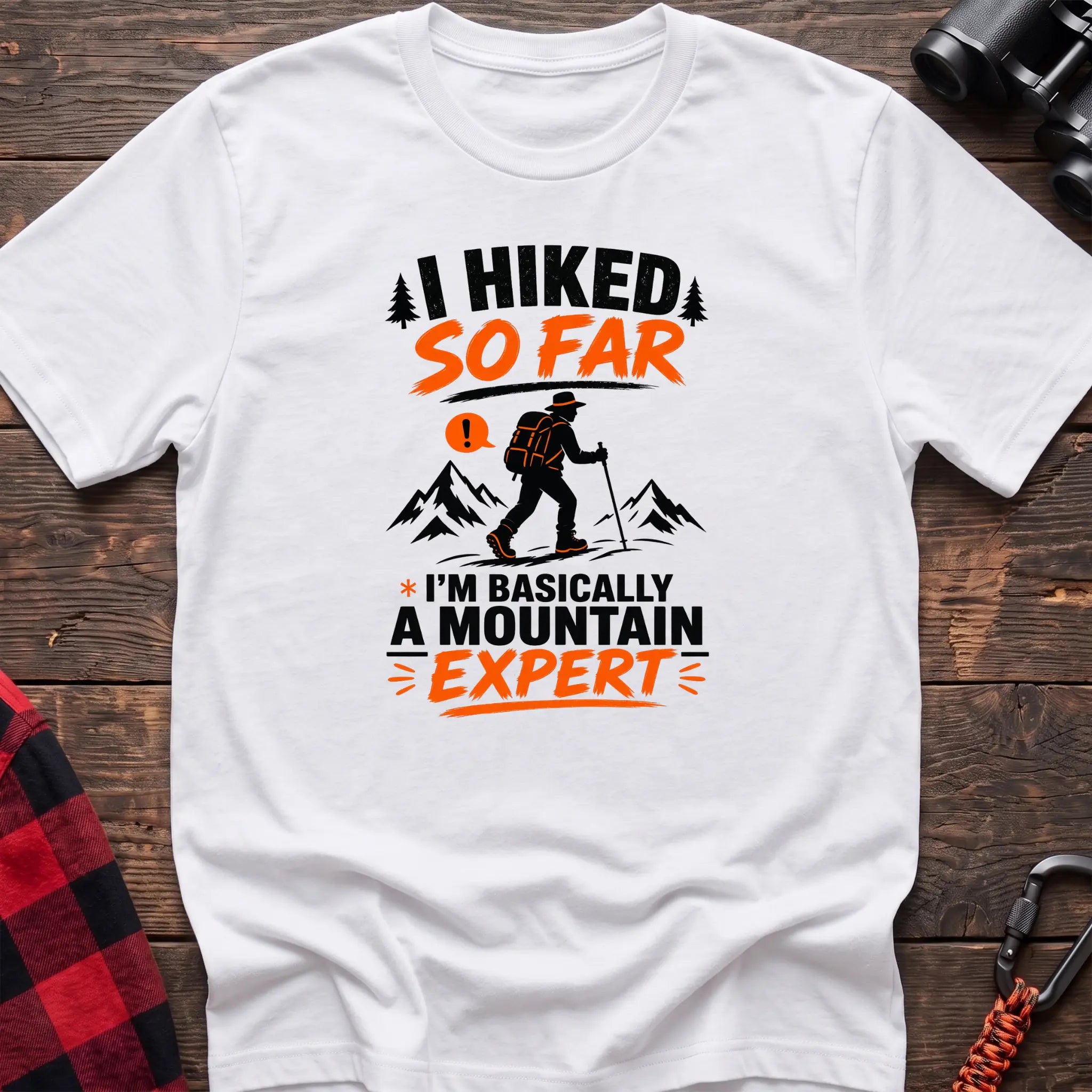 White cotton t-shirt with I Hiked So Far I'm Basically a Mountain Expert graphic of a hiker with backpack and trekking pole, flat lay on rustic wood with hiking gear